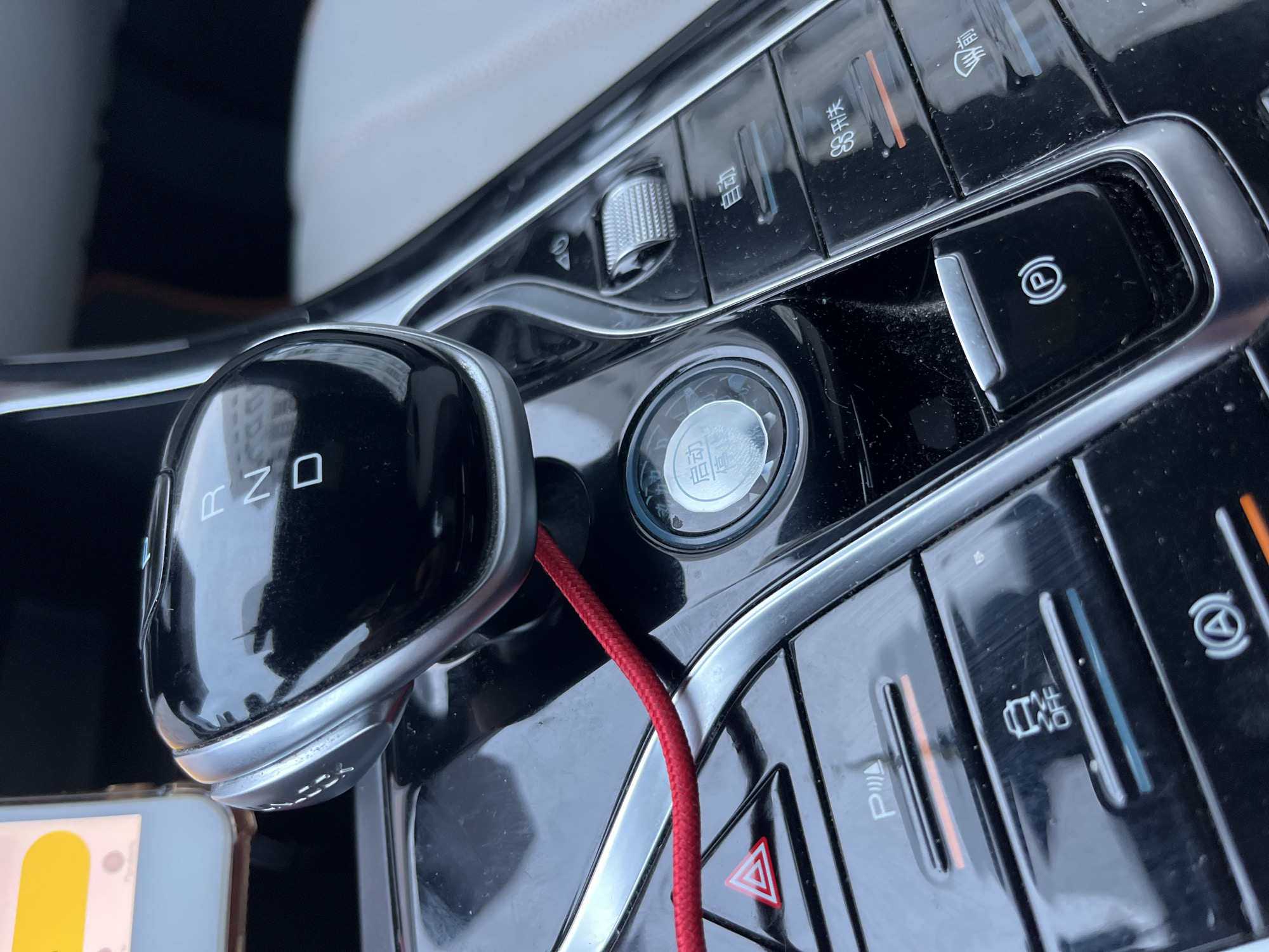 The image shows a close-up view of a car's interior, focusing on the center console. A silver and black gear shifter is prominently displayed, with "R", "N", and "D" labels visible. To the right of the shifter, there is a circular button with Chinese characters that appear to read "启动/停止" (start/stop). Below this button, there are several other car controls, including buttons for parking assist, traction control, and a parking brake. A red braided cord runs across the console. The overall impression is of a modern car interior. The specific location within the city of Minsk, Belarus cannot be determined from the provided image.