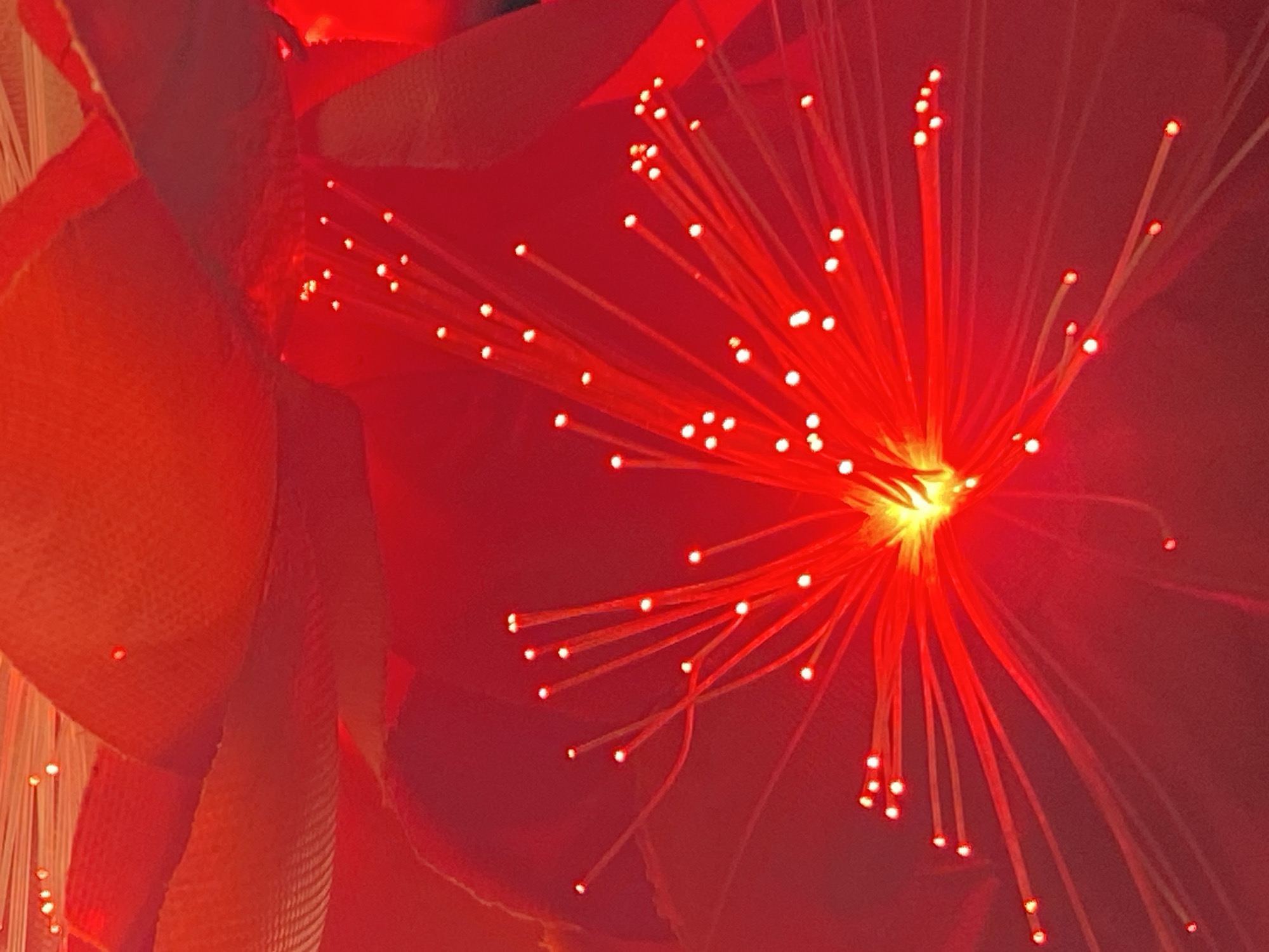 This is a close-up, high-angle shot of a red fiber optic lamp that appears to be shaped like a flower. The light source is at the center, emanating thin red fiber optic strands with small, bright white lights at the tips. The background consists of soft red petals or fabric. There is no visible indication of the location in Abakaliki, Nigeria, or any people or specific actions.