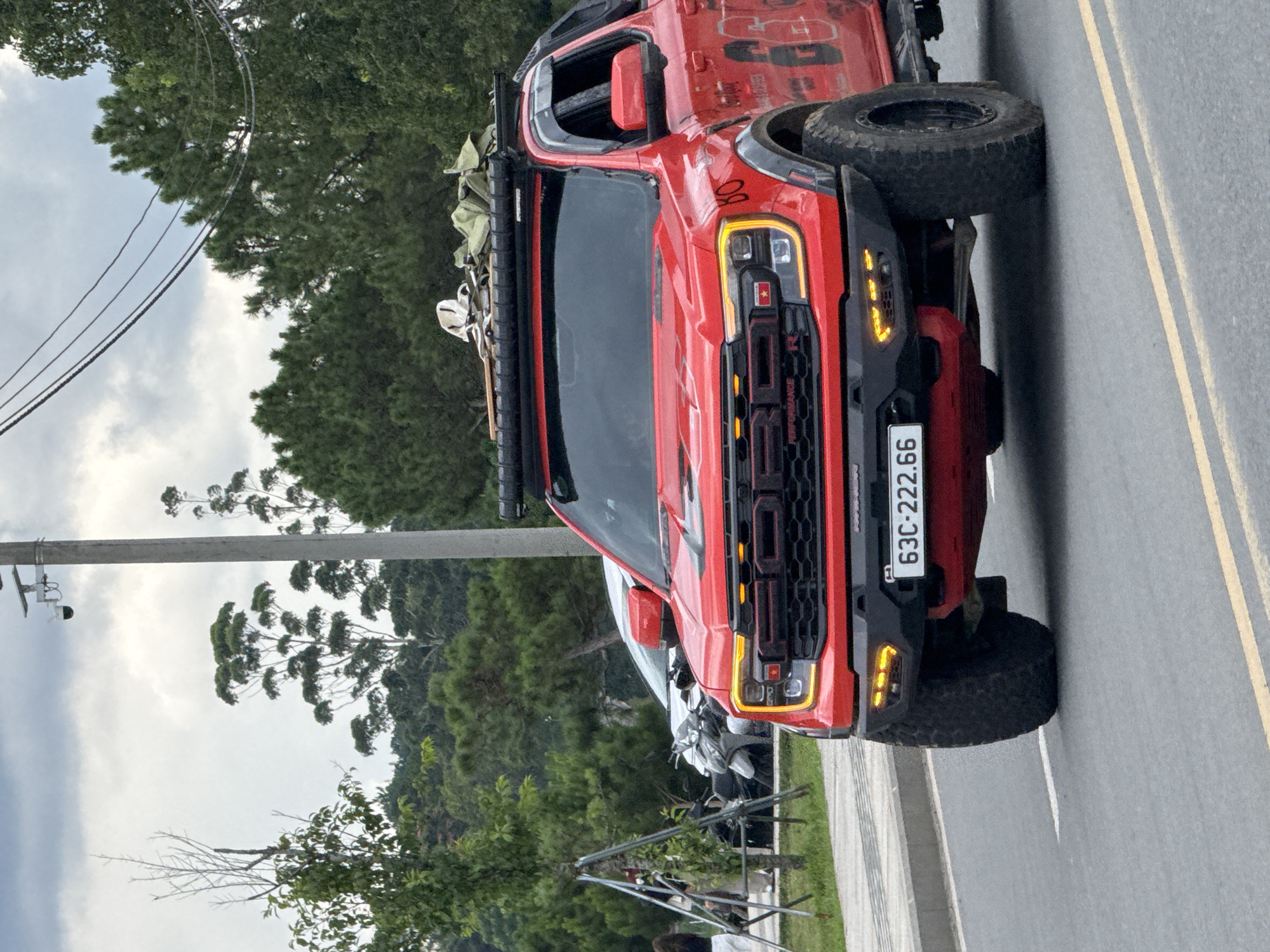 A red Ford Raptor pickup truck is driving on a paved road in Da Lat, Vietnam. The truck has a roof rack loaded with cargo and a license plate that reads "63C-222.66". In the background, there are trees, a white car, and motorcycles. The sky is overcast. A security camera is visible on a pole on the left side of the image.