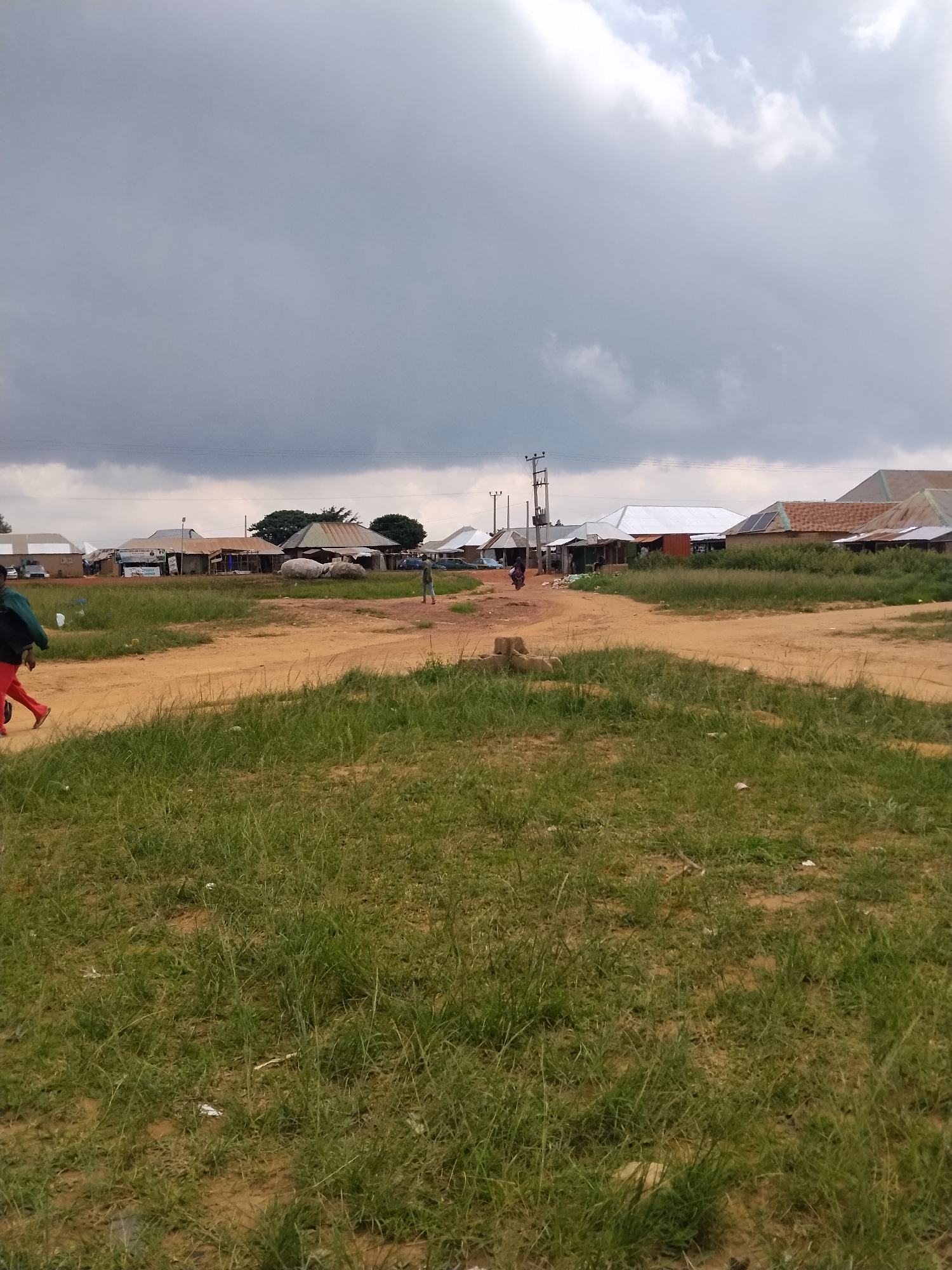 The image depicts a street scene in Durumi, Nigeria, characterized by a dirt road with patches of green grass on either side. In the background, a collection of buildings with corrugated metal roofs are visible, suggesting a residential or commercial area. The sky is overcast with dark, heavy clouds, hinting at impending rain. Several individuals are present, with some walking on the road and others seemingly engaged in activities near the buildings. The overall impression is one of a typical, unpaved urban environment under a moody sky.