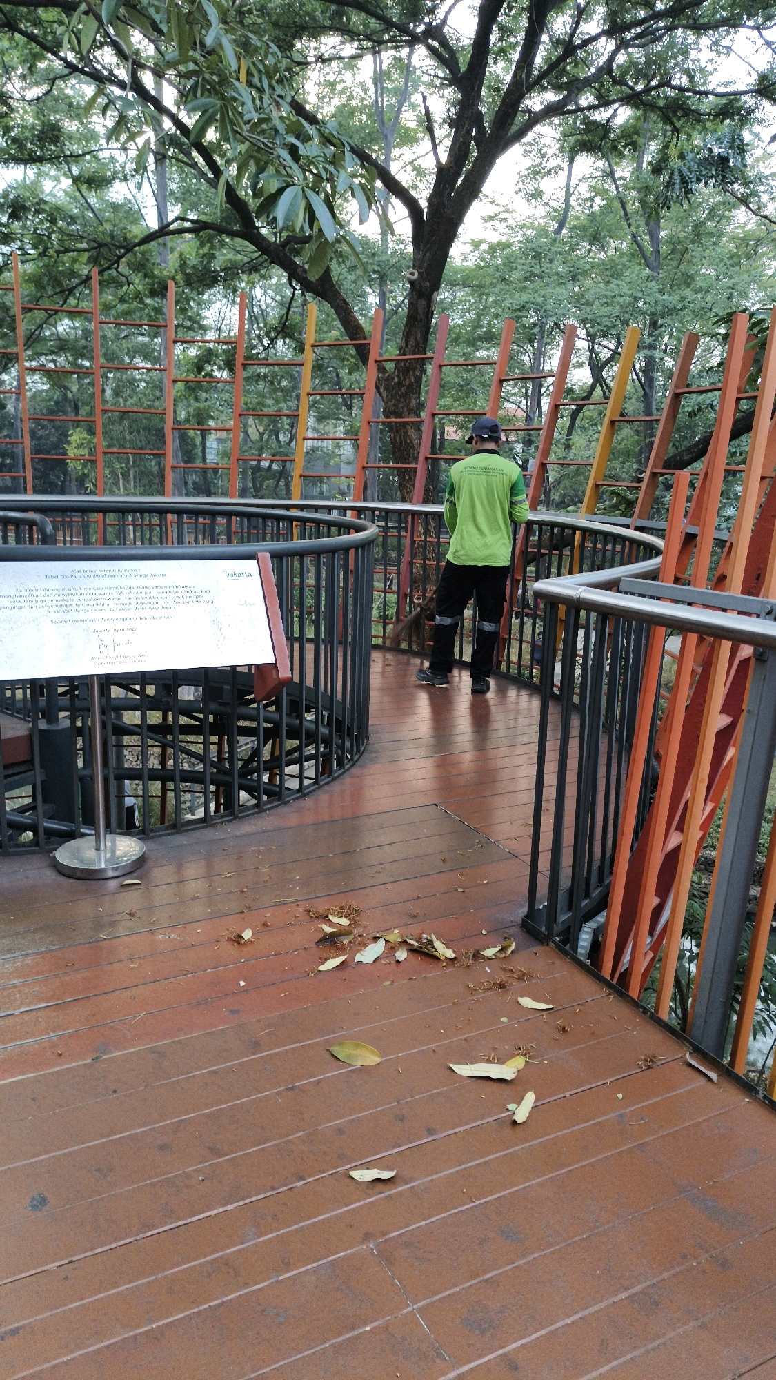 In South Jakarta, Indonesia, a person in a green long-sleeved shirt and black pants, wearing a dark baseball cap, is sweeping a wooden walkway with a broom. The walkway curves to the right, with a metal railing along its edge. Behind the walkway, a spiral staircase can be seen descending. The area is surrounded by trees and an orange and yellow structure with vertical slats. A sign is mounted on a metal pole to the left of the walkway. Fallen leaves are scattered across the wooden planks. The lighting suggests it is daytime, possibly late afternoon.