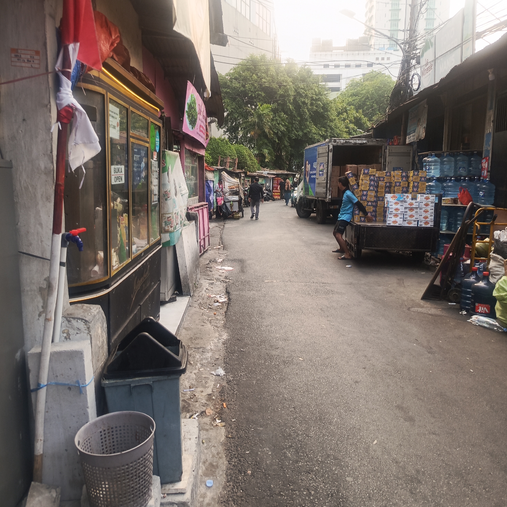 The image depicts a street scene in a commercial area, likely a market or a row of shops, in South Jakarta, Indonesia. The foreground shows a concrete sidewalk with a large grey trash bin and a metal mesh waste bin next to it. A red and blue faucet is attached to a pipe on a concrete pillar. The street is paved and appears to be a one-way road with vehicles parked or moving along it.
On the left side of the street, a building with large glass windows is visible, displaying various items. A red and white Indonesian flag is draped over a pole in front of this building. Further down the street, several small shops or stalls with corrugated metal roofs line the road.
On the right side of the street, a truck is parked, partially loaded with boxes and large water cooler jugs. A person wearing a blue shirt and black shorts is seen near the back of the truck, interacting with the cargo. The background shows dense green trees and modern high-rise buildings, suggesting a blend of older commercial structures and newer developments within the city. The sky is overcast, giving the scene a somewhat muted lighting. Scattered litter is present on the pavement. Several individuals are walking along the street in the distance.