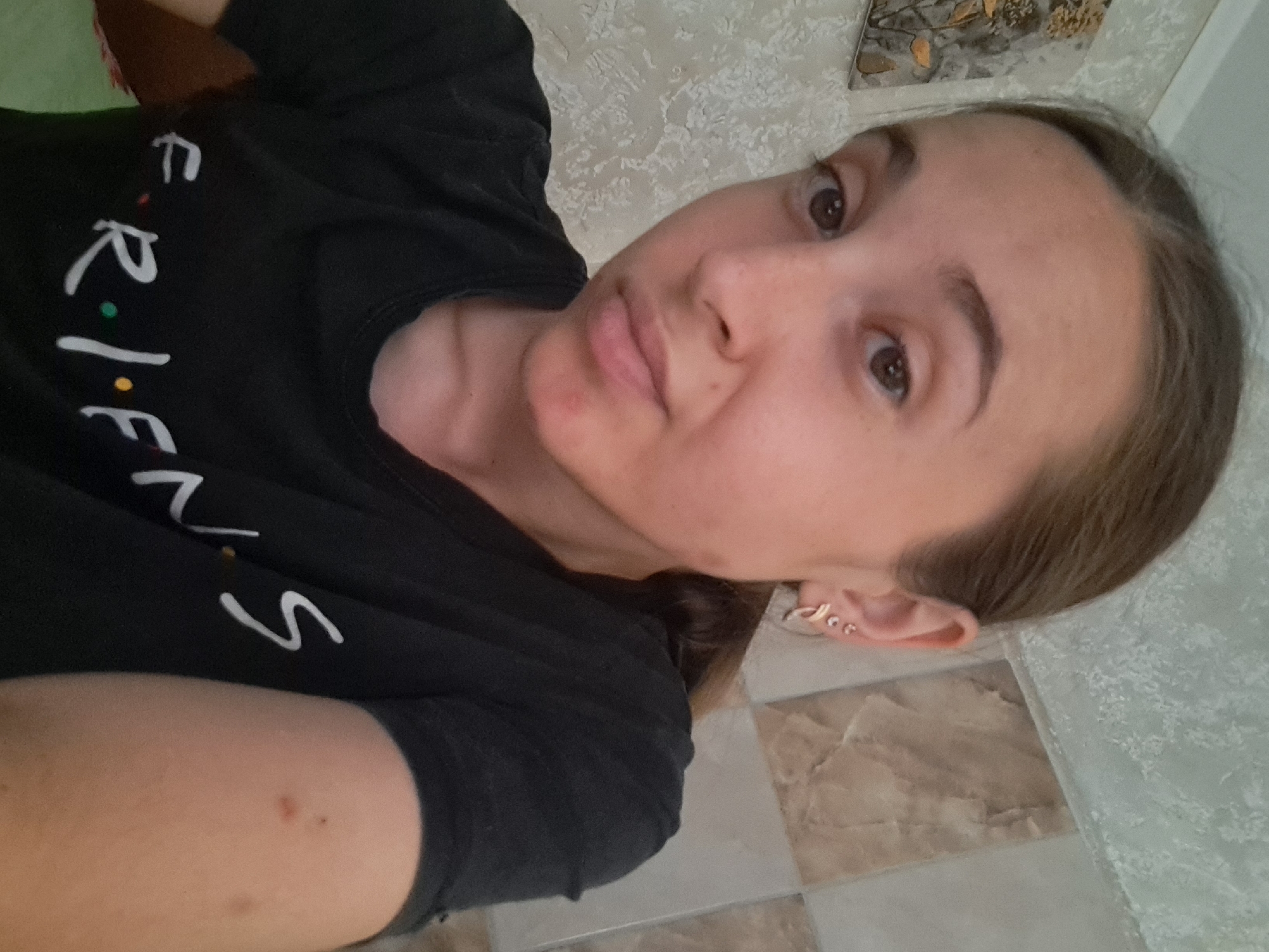 A young woman with brown hair pulled back and wearing a black "Friends" t-shirt is pictured in a close-up, selfie-style shot. She has brown eyes and is wearing hoop earrings. The background appears to be a wall with a textured wallpaper and some tiled sections, suggesting an indoor residential setting. The lighting is natural, casting a slight shadow on her face. Her left arm, visible on the right side of the frame, has a few small blemishes. The image quality is slightly grainy, typical of a smartphone photo taken indoors. There is no specific indication of her location within Vypasne, Ukraine.