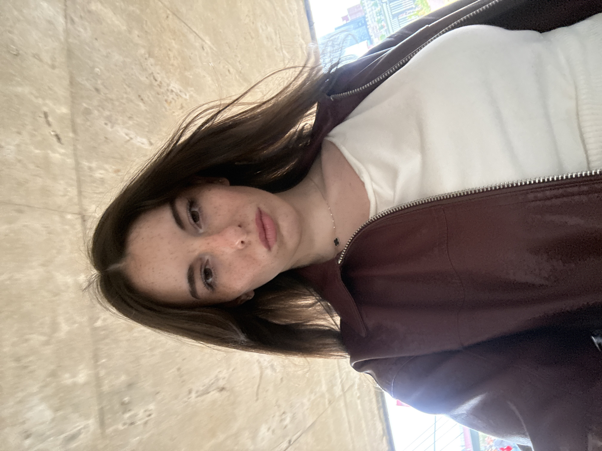 A young woman with dark brown hair and freckles is pictured in a selfie. She is wearing a maroon leather jacket over a white ribbed top and a delicate silver necklace with a black pendant. Her hair is windswept and frames her face. The background is a textured, light-colored ceiling or overhang, possibly concrete, with some subtle patterns. In the distance, through an opening or gap, city buildings are visible, suggesting an urban setting. It is not possible to definitively determine the specific location within Rotterdam, Netherlands, from the provided image.