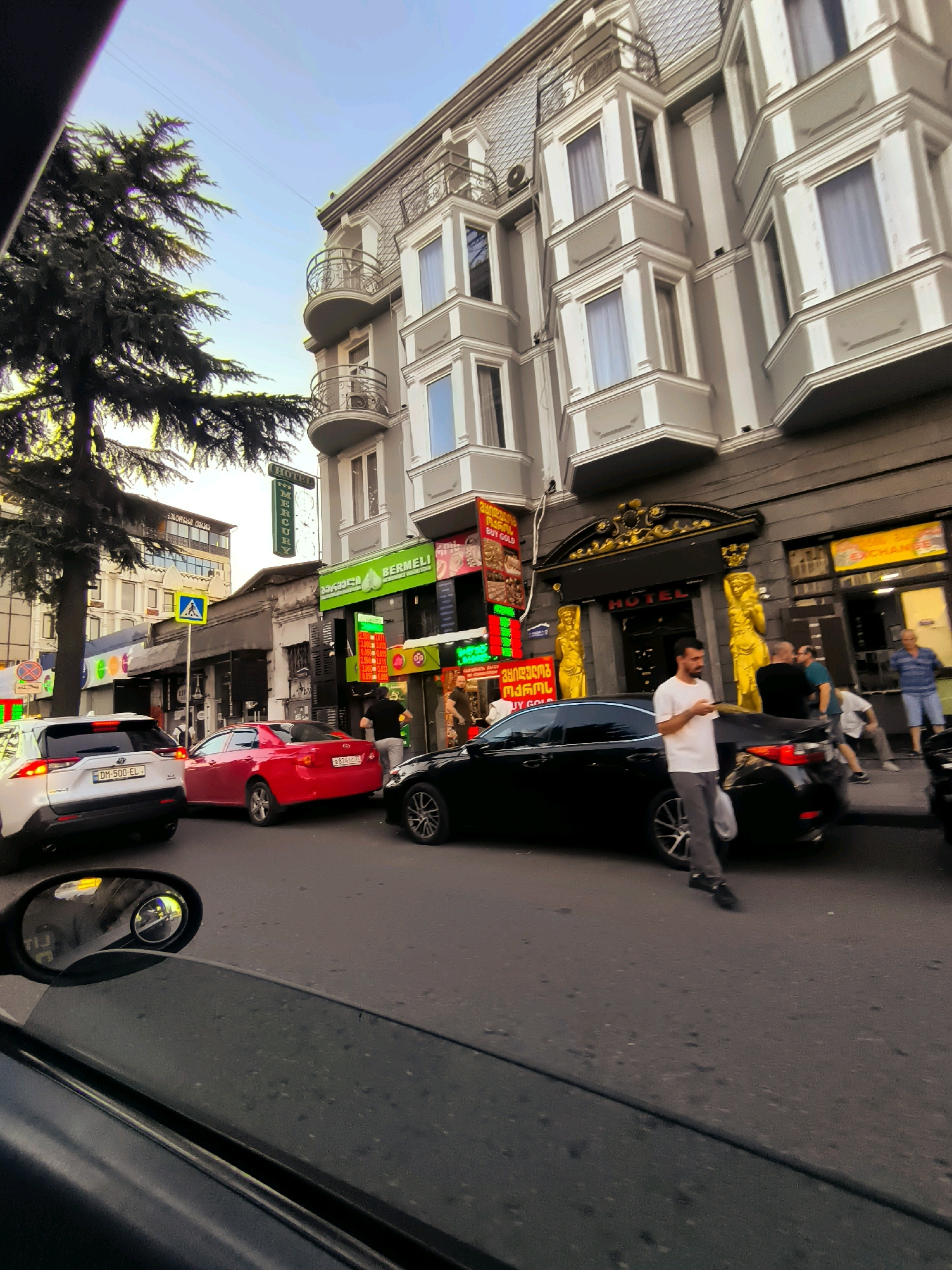 In an urban street scene, a white SUV is parked in the foreground, its rear facing the viewer. To its left, a red sedan is seen. Beyond these vehicles, a black sedan is parked at the curb. A man in a white t-shirt and gray pants stands next to the black sedan, looking at his phone. Several other individuals are visible on the sidewalk and street in the background. The buildings lining the street feature multiple stories with ornate balconies and windows. Signs for "HOTEL," "BERMELI," and "BUY GOLD" are displayed on the buildings. A tall evergreen tree is situated on the left side of the frame. The sky above is clear and blue. A car side mirror in the lower left corner of the image reflects the street scene, including a yellow traffic light.