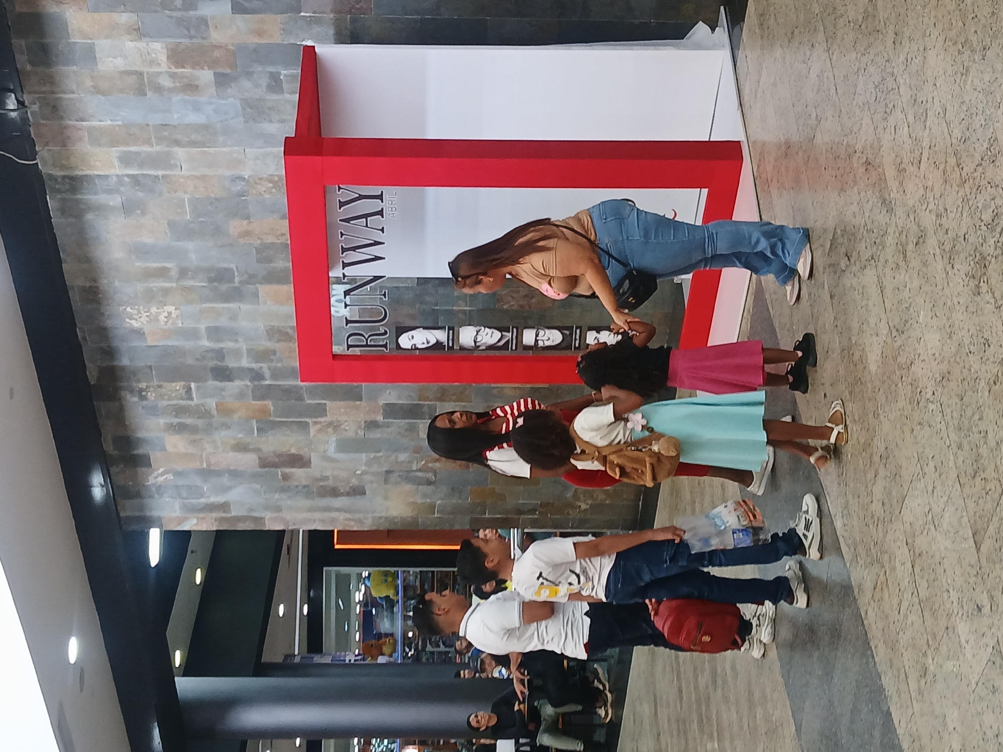The image captures an indoor scene, likely a shopping mall or public exhibition space, in Ciudad Guayana, Venezuela. The primary focus is a red-framed display positioned against a gray brick-patterned wall, with a polished concrete floor in the foreground.
At the display, a woman with long brown hair, wearing a beige top and blue jeans, is leaning forward, seemingly interacting with the exhibit or posing for a picture. The display prominently features the word "RUNWAY" written vertically in black letters, with "ABRIL" (Spanish for April) beside it. Below "RUNWAY," there are five black and white portraits of faces arranged horizontally, suggesting a fashion-related event or promotion.
Several other individuals are present in the vicinity. To the right of the bending woman, a child in a pink skirt stands next to an adult wearing a light blue dress and carrying a brown bag. Another adult, dressed in a red and white striped top and red skirt, stands nearby. In the lower left quadrant, a group of three men are seen. One man in a white t-shirt and jeans carries a red backpack, while another in a white t-shirt and jeans holds a plastic bottle. The background shows parts of the building's interior, including black support beams and bright overhead lights. The overall atmosphere appears casual, with people observing or casually passing by the display during what seems to be daytime.