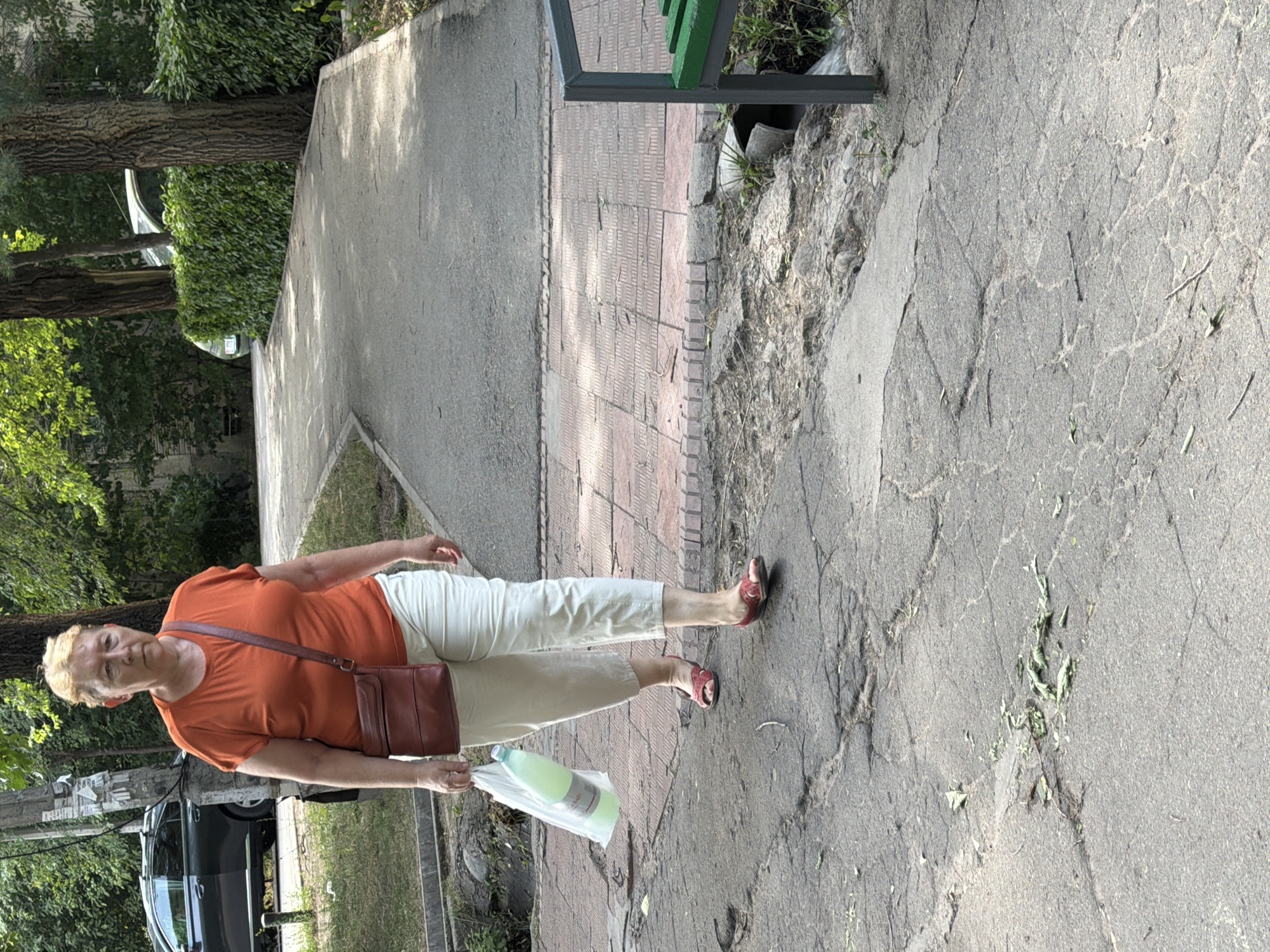 A woman with short blonde hair, wearing an orange short-sleeved shirt and light-colored pants, walks on a cracked asphalt path. She carries a large clear plastic bag containing a light green bottle in her left hand and has a brown crossbody purse on her right shoulder. She is wearing red sandals. The path is bordered by a red brick walkway to her left and appears to be in an urban park or residential area with trees and foliage visible in the background. A dark-colored car is parked on the left side of the frame. The scene is illuminated by daylight, casting shadows.