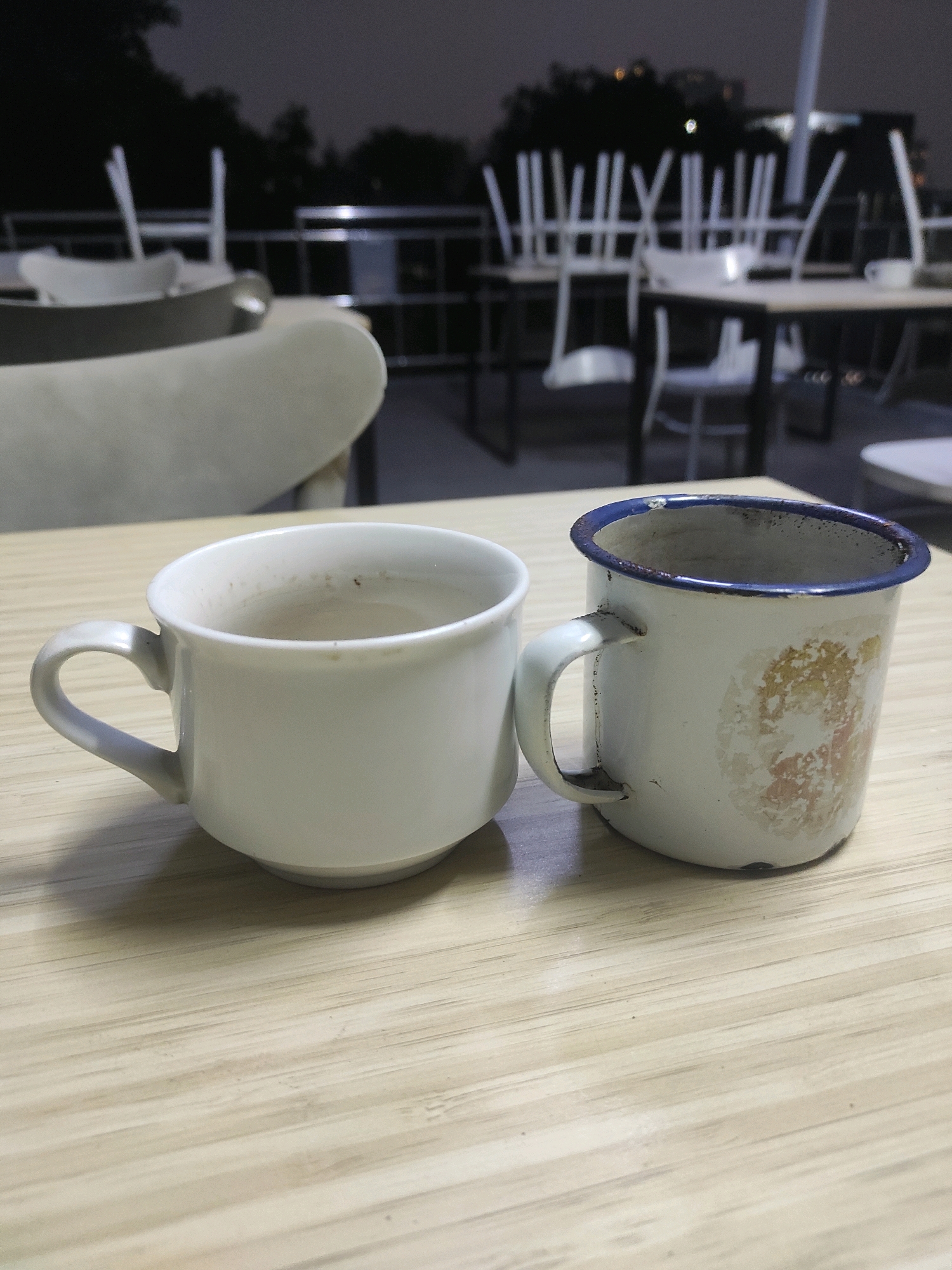 The image shows two cups on a wooden table. One cup is white and appears to be ceramic, with a subtle sheen. The other cup is white enamel with a blue rim and some wear and tear, including a discolored area with faint yellow and orange markings. The background is blurred, showing outdoor seating with stacked chairs and tables, suggesting a cafe or restaurant setting, possibly at night due to the dim lighting and dark sky. The overall scene is still and quiet.