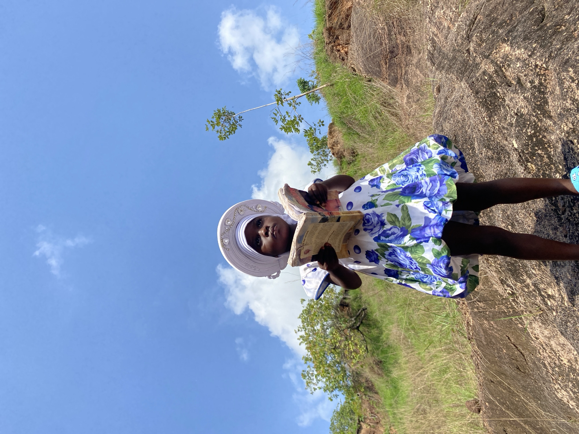 A young girl, likely of African descent, stands outdoors in a natural, sunlit environment. She is positioned slightly off-center to the left, looking directly at the viewer with a neutral, perhaps contemplative, expression.
The girl is dressed in a white short-sleeved dress adorned with a prominent blue and green floral pattern, primarily depicting roses. On her head, she wears an elaborate white head covering, possibly a hat or headwrap, featuring decorative embellishments that appear to be small white beads or sequins. Her visible leg is dark-skinned, and she is wearing light blue or turquoise shoes. She holds an open book or magazine in both hands, positioned in front of her. While the text on the pages is largely illegible due to the angle and resolution, faint words like "MOU R," "HAIL," "LORD," and "GOD" can be partially discerned, suggesting the content might be religious or literary.
The setting is an outdoor, hilly landscape. The background features a bright, clear blue sky dotted with scattered white clouds, indicating sunny weather. Below the sky, several small trees or shrubs with green foliage are visible. To the right of the girl and extending behind her is a steep, rocky incline or hillside, partially covered with patches of dry, golden-brown grass and some greener vegetation. The bright natural light suggests it is daytime, likely late morning or early afternoon. The scene evokes a tranquil outdoor portrait.