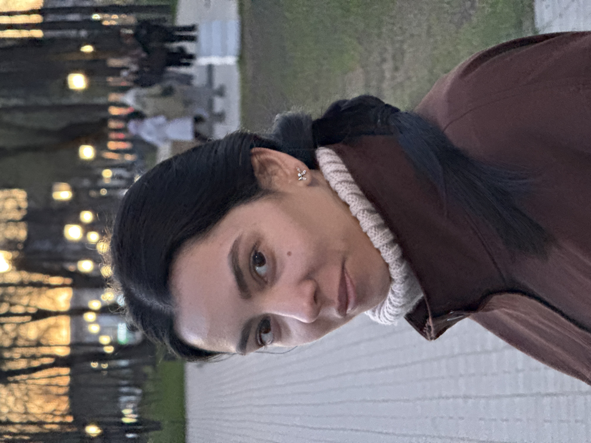 A woman with dark hair and brown eyes is pictured in an outdoor setting. She's wearing a brown jacket over a cream-colored turtleneck sweater. Her hair is pulled back, revealing a small, flower-shaped earring in her left ear. The background appears to be a park or urban area with lights and trees visible in the blurred distance. The ground is paved with a light-colored material. There is no specific indication that the image was taken in Chaikovsky, Russia.