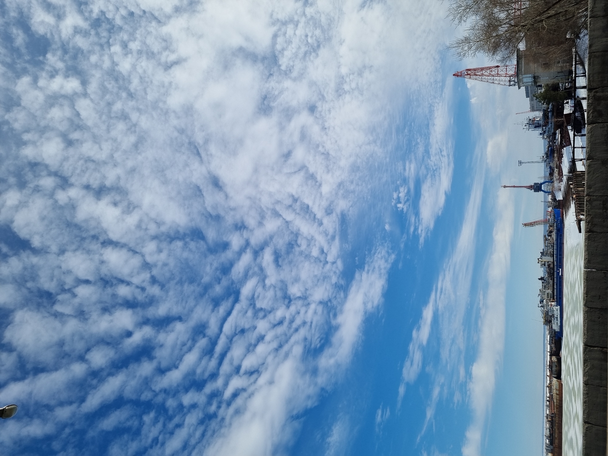 The image depicts a port or shipyard facility located in Chaikovsky, Russia, likely situated on the Kama River or the Votkinsk Reservoir. The scene is observed from an elevated position, looking out over a concrete barrier that runs across the lower foreground. The sky is a clear blue, approximately 70% covered by white altocumulus clouds arranged in a distinct ripple pattern. A partial view of a street lamp fixture is visible in the upper left corner of the frame.
The mid-ground features a body of water largely covered with broken ice and slush. Numerous vessels, including several visibly rusty barges and other ships of various sizes, are moored or docked within the icy expanse. In the background, an industrial port complex is visible, characterized by multiple large cranes. A prominent red-and-white lattice boom crane with an attached control booth stands on the far right. Further to the left, a large blue gantry crane is visible, along with several other smaller cranes and industrial structures. One multi-story building is situated adjacent to the red crane. Patches of snow are evident on the ground within the port area. Bare deciduous trees or shrubs are present on the far right side of the image. No individuals are discernible within the scene.