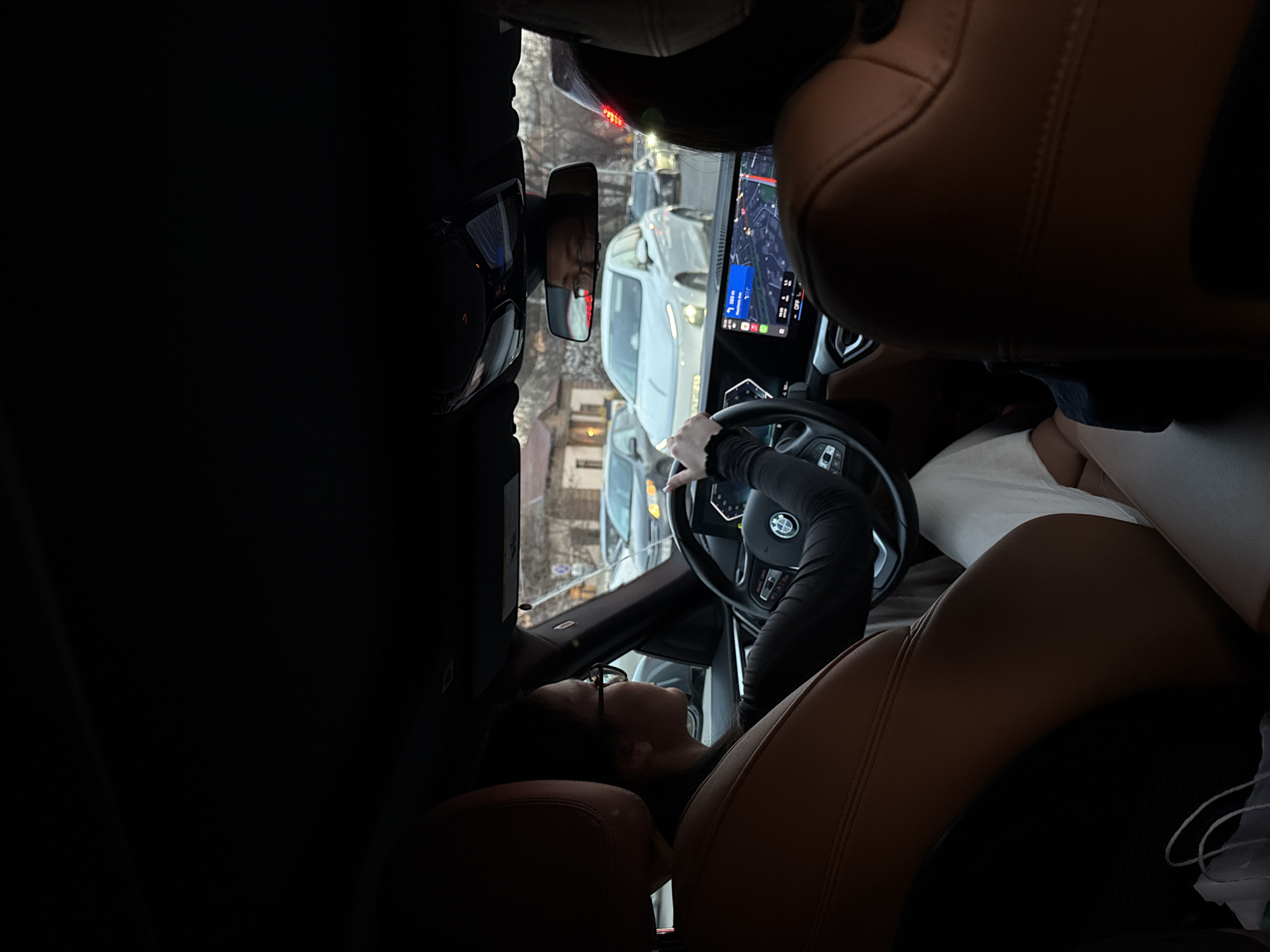 A person with glasses is seated in the driver's seat of a car and is looking upward. The person is gripping the steering wheel. The car's navigation screen is displaying a map, and a rearview mirror reflects the surrounding street, which shows other cars and buildings. The location appears to be a city street, and is, based on the navigation, in Yerevan, Armenia. The interior of the car features tan leather seats.