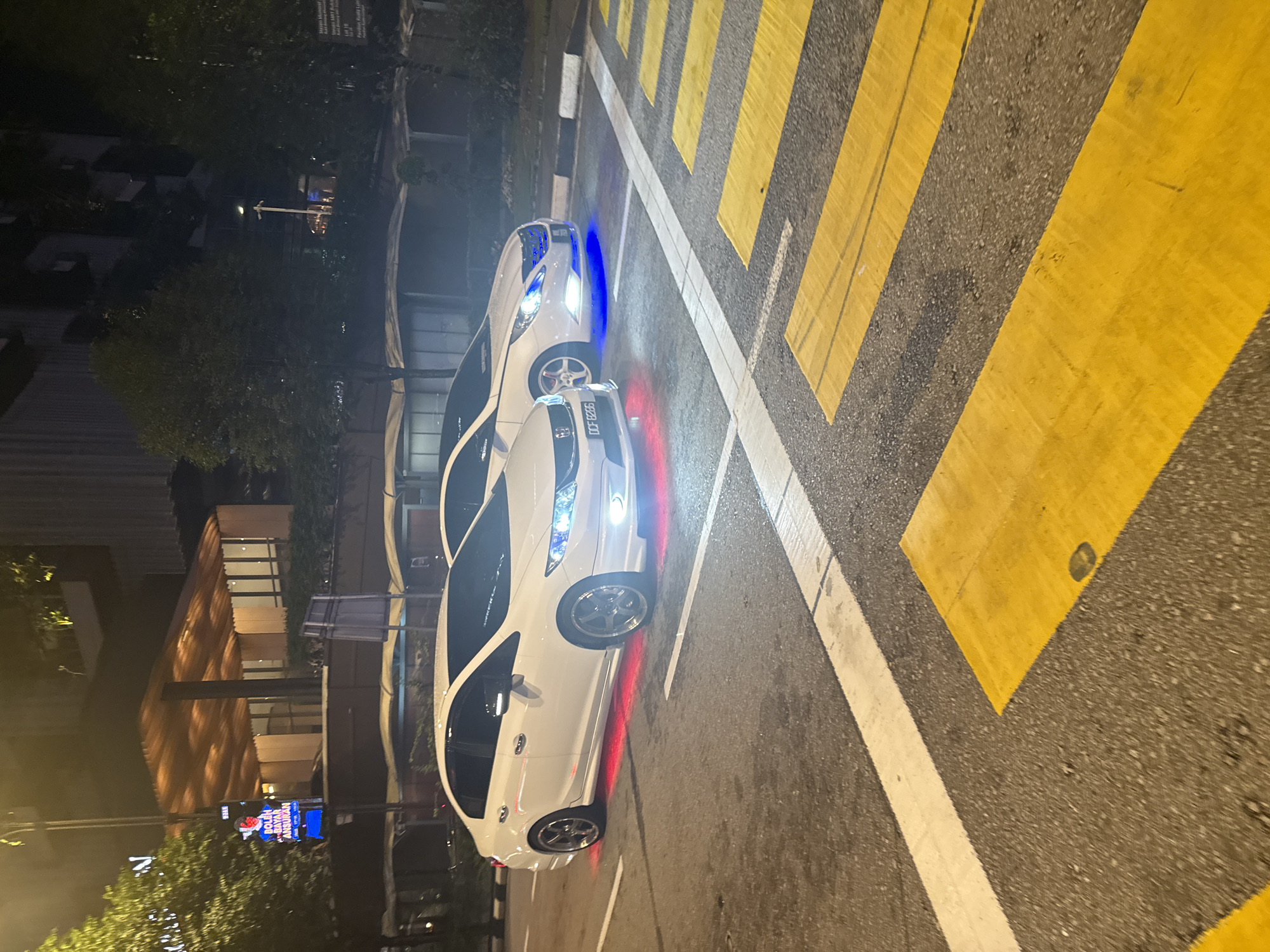The image captures a nighttime urban scene in Kuala Lumpur, Malaysia, featuring two white cars prominently displayed on a road with distinctive markings. The cars, both appearing to be sedans, are stationary. The front car exhibits bright white headlights and custom red underglow lighting, illuminating the damp asphalt beneath it. Its license plate, "DCF 8286," is clearly visible, and it features aftermarket silver multi-spoke rims. Behind it, a second white car is visible, distinguished by blue underglow lighting and blue-tinted headlights or grille accents.
The cars are positioned on a road marked with a broad yellow and black zebra crossing, along with white solid and dashed lines, indicating a pedestrian crossing or intersection. A black and white striped kerb runs along the side of the road.
In the background, various urban elements are visible: multi-story buildings, some illuminated, and several trees. A street sign with an arrow is faintly discernible, and a billboard with glowing blue text reads "BOLEH BAYAR ANSURAN" (Malay for "Can pay installments"), alongside a prominent 'N' logo. The overall setting is dark, suggesting late evening or night, with artificial lights providing illumination to the scene. The road surface appears damp, possibly from recent rain or high humidity.