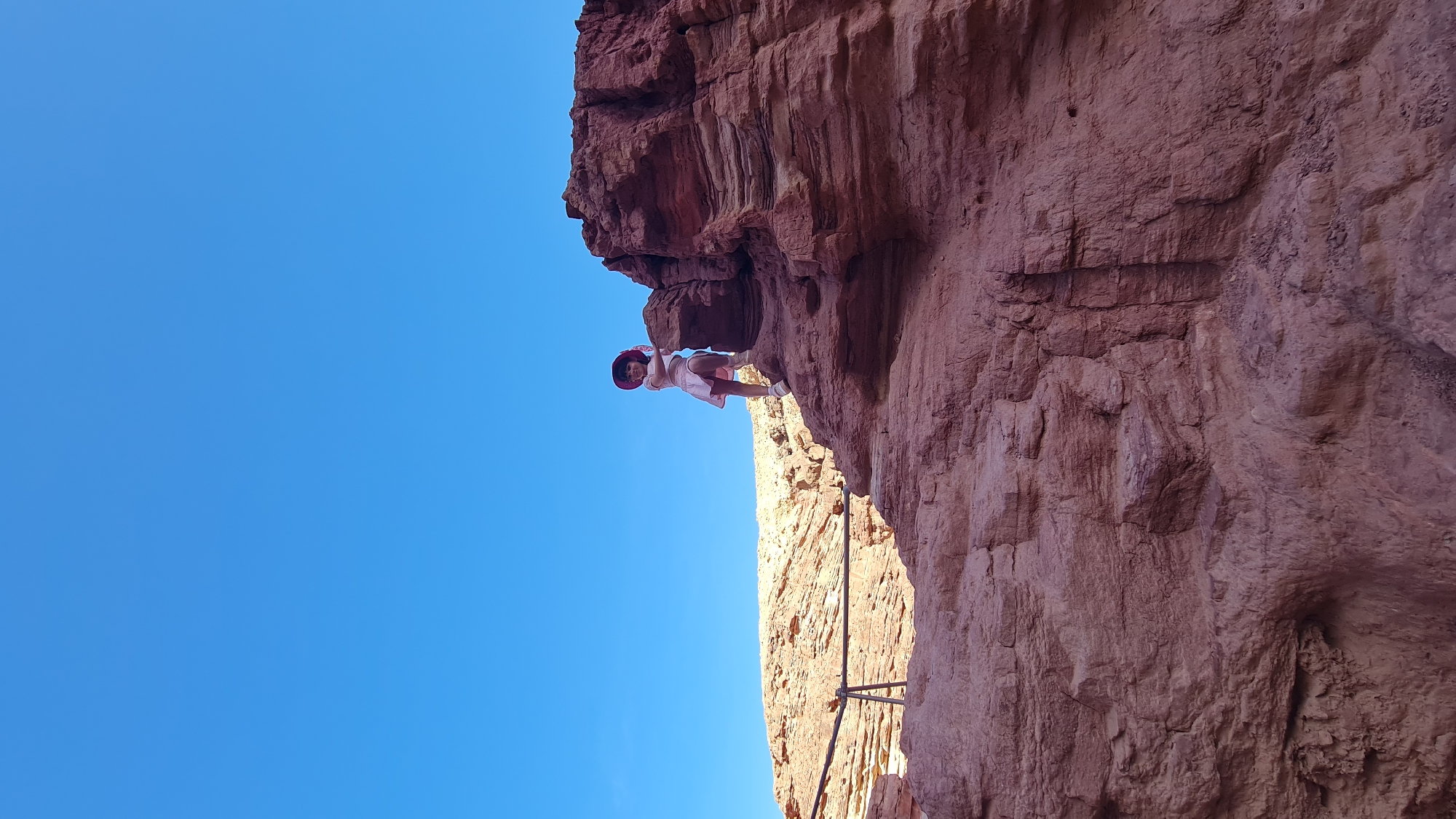 A girl stands on the edge of a red rock formation under a clear blue sky in Hevel Eilot Regional Council, Israel. The rock formation is layered and textured, with a handrail visible below the girl. She is wearing a light-colored dress and a hat.