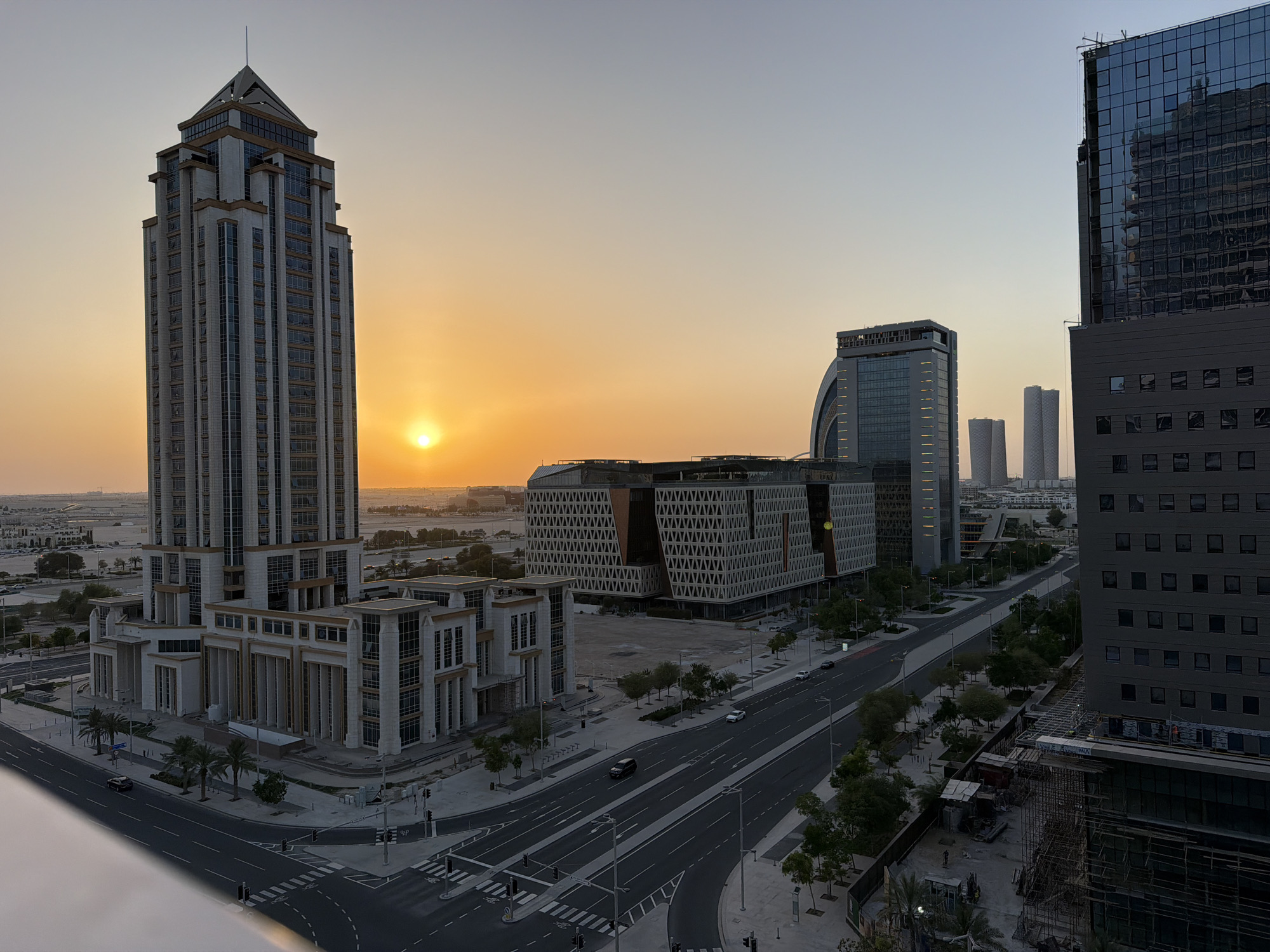 An aerial view captures a city skyline at sunset. The sun, a bright orange orb, is low in the sky, casting a warm glow over the scene. Dominating the left side of the image is a tall, multi-story skyscraper with a pyramidal roof. The building's facade is a combination of white stone and glass windows, with decorative golden accents. In front of the skyscraper is a lower, wider building with a similar architectural style.
To the right of the skyscraper, several other modern buildings rise. One features a distinctive curved, arched glass facade, and another is a tall, dark glass skyscraper reflecting the sky and surrounding structures. In the mid-ground, a large, modern building with a patterned geometric facade stands out. Roads with clear lane markings and streetlights are visible, with a few cars driving on them. Palm trees and other greenery line the streets and sidewalks. The overall environment suggests a modern urban setting. There are no people clearly visible in the image.