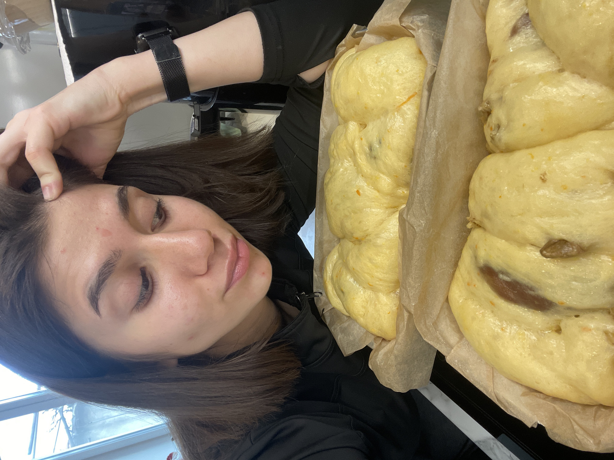 A woman with shoulder-length brown hair, wearing a black long-sleeved shirt and a black watch, looks down at the camera while touching her hair. To the right of the woman are two loaves of braided challah bread sitting in baking paper within what appears to be a black baking pan. The background is somewhat blurry, but a window and a portion of a kitchen or countertop are visible. There is no clear indication of this being in Sofia, Bulgaria.