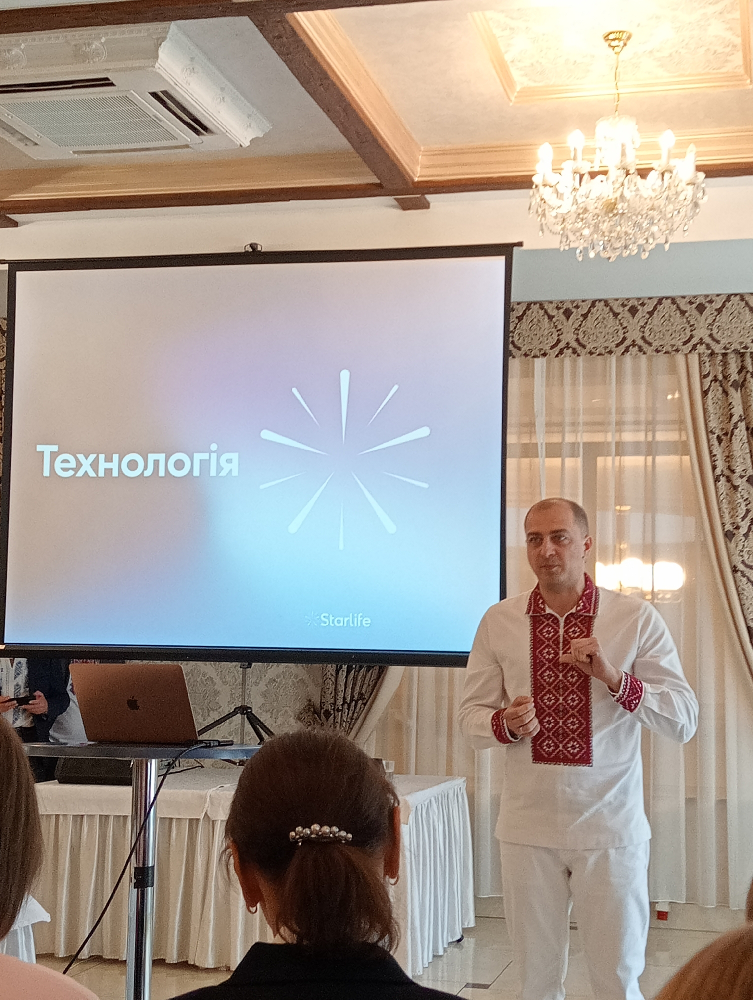 The image depicts a presentation or seminar in Lviv, Ukraine, featuring a man standing in front of a projection screen. The screen displays the word "Технологія" (Technology) along with a starburst graphic and the name "Starlife". The man is wearing a white shirt and pants, adorned with traditional Ukrainian red embroidered patterns. He is gesturing with his hands as if speaking. A laptop sits on a table in front of the screen. In the foreground, the back of a woman's head is visible. The environment includes a chandelier and patterned curtains. The room suggests a formal or upscale event space.