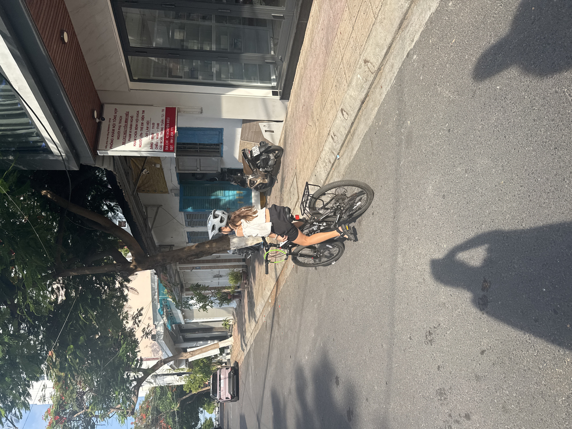 A young girl wearing a white helmet and a white top with black shorts rides a bicycle on a sunny paved street in Vietnam. She is positioned in the middle of the frame, with the bicycle in motion. The background shows a row of buildings with storefronts and windows, and some trees with green foliage and red flowers. A sign with Vietnamese text is visible on one of the buildings. The street is made of asphalt, and there is a sidewalk made of beige tiles. The shadow of a person holding a camera is cast on the street to the right of the frame, indicating the photo was taken by someone. The lighting suggests it is daytime, likely late morning or afternoon due to the strong shadows. A parked motorcycle and a car are also visible further down the street. The overall scene depicts a casual, everyday moment in a bustling urban environment. The visible text is a sign for a medical clinic, listing its name, doctor's name, services, working hours, address, and phone number.