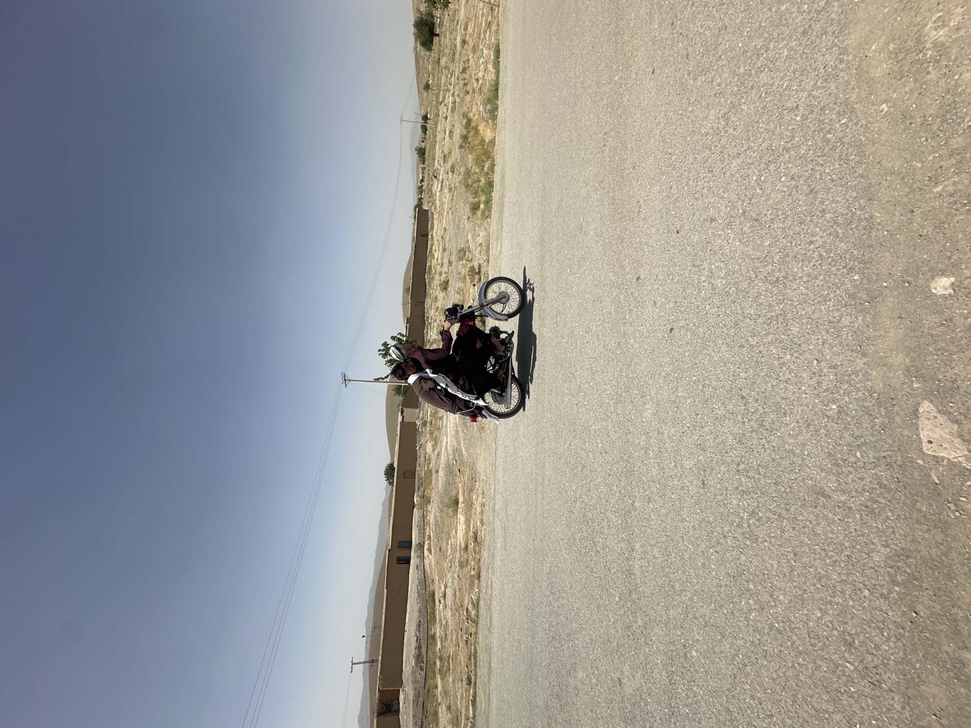 Two individuals, one driving and one riding as a passenger, are seated on a blue and silver motorcycle traveling on an asphalt road. The driver, on the left, wears a dark brown outfit and a white turban. The passenger, on the right, is also dressed in dark clothing and wears a white head covering. They are both facing forward, looking in the direction of travel.
The motorcycle appears to be moving at a moderate speed. The background depicts a dry, arid landscape with a long, light brown building or wall stretching horizontally across the frame. Beyond the wall, rolling hills are visible under a clear, pale blue sky. Utility poles and power lines are present in the background, indicating infrastructure in the area. The road surface is asphalt with some gravel and debris scattered along the edges. The lighting suggests it is daytime, with the sun casting a bright light, creating a slight haze in the distance. The location is identified as Kanozai, Pakistan.