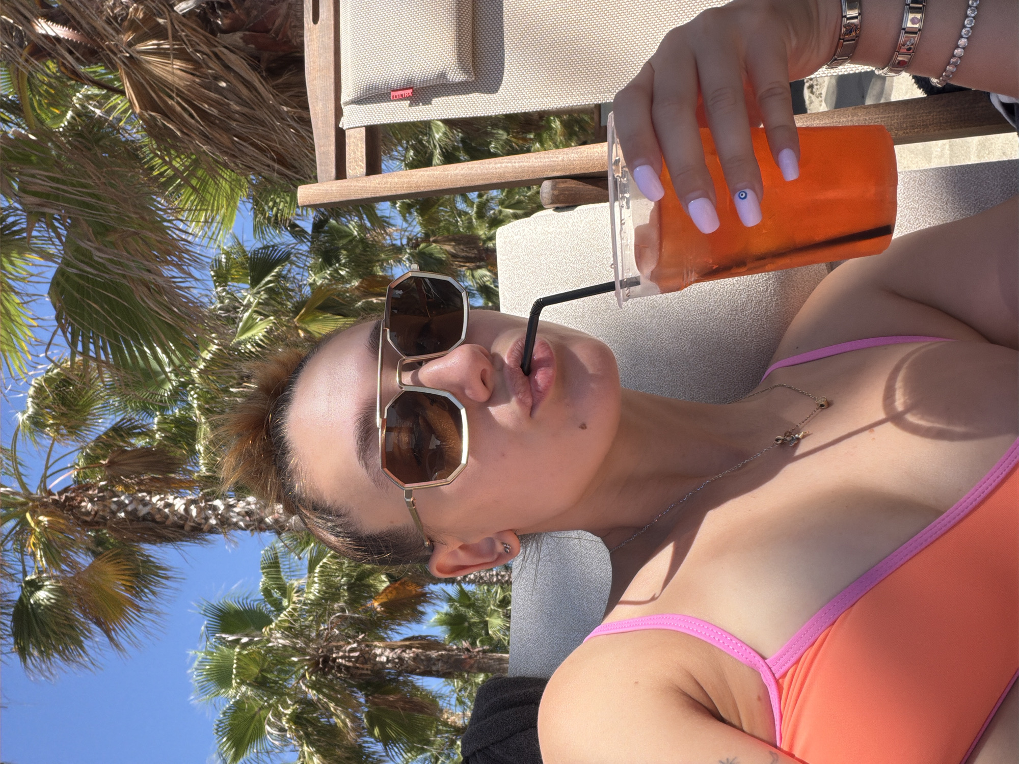 The image features a woman lounging on a chair, holding a clear plastic cup filled with an orange beverage and a black straw. She is wearing sunglasses with gold frames and a coral bikini with pink trim. Her nails are painted a light lavender color, and she has a delicate silver necklace with a small pendant. Her right hand, adorned with bracelets, is near the cup. The background shows lush green palm trees against a clear blue sky, suggesting a warm, sunny day. The woman's face is close to the camera, and she appears to be taking a sip of her drink, with her lips slightly pursed around the straw. The overall impression is one of relaxation and enjoyment in a tropical or Mediterranean setting. A tag visible on the chair in the background reads "ELEKTRA".
