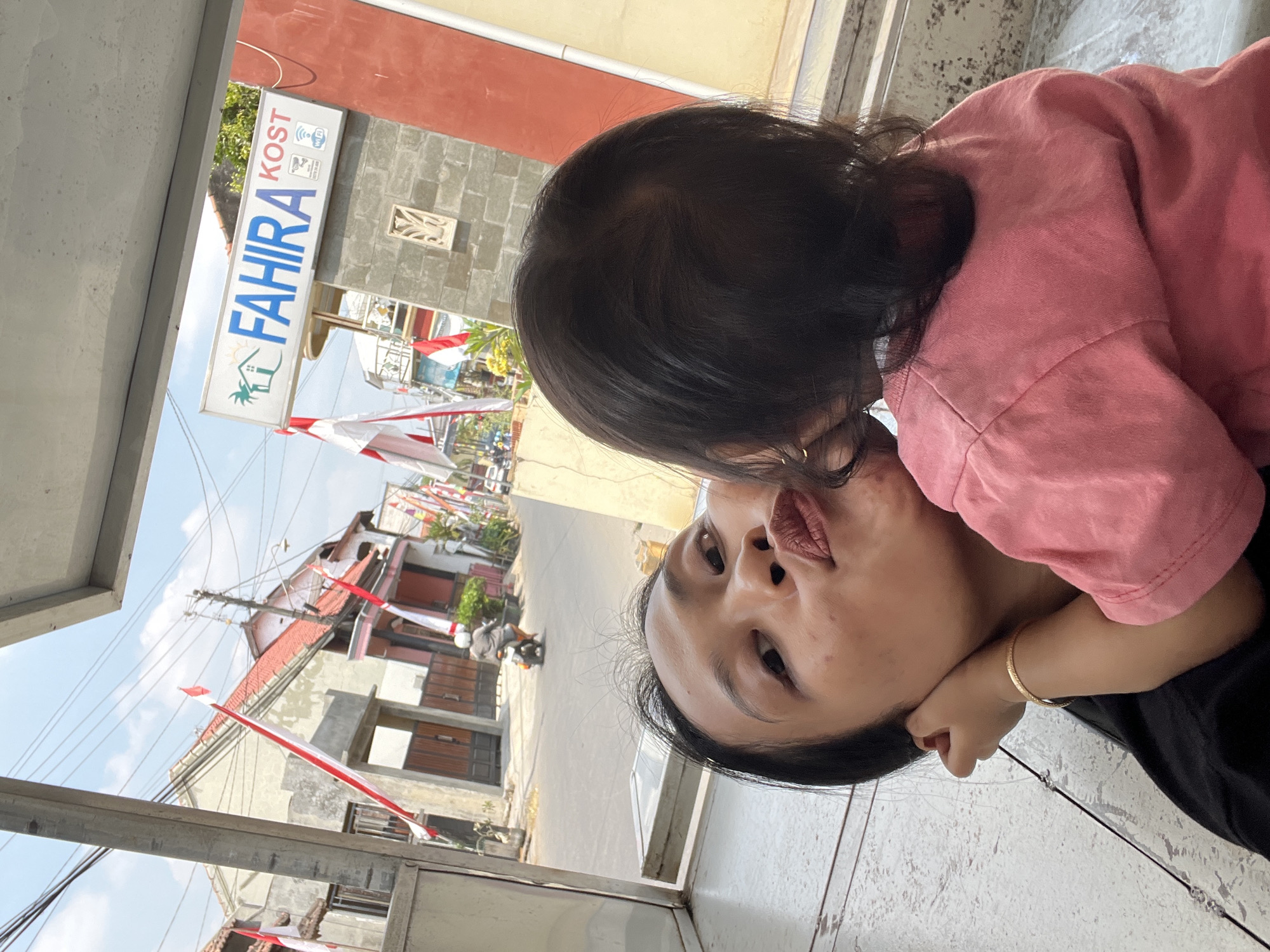 In the city of Kecamatan Pati, Indonesia, a woman and a young child are captured in a selfie taken outdoors. The woman, positioned in the foreground on the left, leans in for a kiss from the child. She has dark hair and is wearing a black top. The child, facing away from the camera and towards the woman, is wearing a pink top. Behind them, a street scene unfolds with buildings adorned with Indonesian flags, suggesting a national holiday celebration. A sign for "FAHIRA KOST" is visible on a building in the background. The environment is bright and sunny, with a clear blue sky.