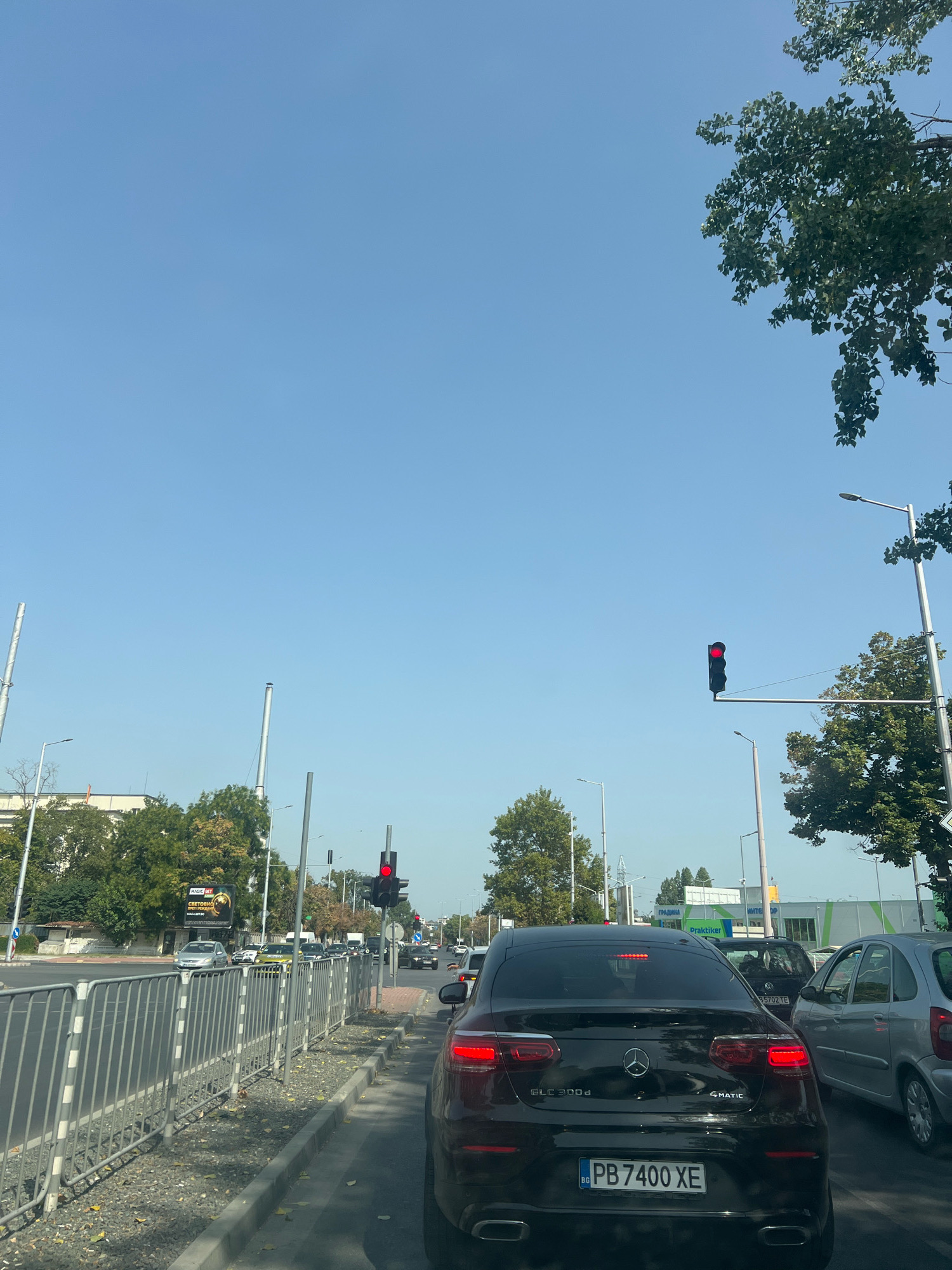 The image shows a street scene in Plovdiv, Bulgaria, with traffic stopped at a red light. The perspective is from inside a vehicle, looking forward. Several cars are visible, including a black Mercedes-Benz GLC 300d 4MATIC with Bulgarian license plate PB 7400 XE, a silver car, and other vehicles in the background. The traffic light is red. On the right side of the road, there is a metal fence separating the road from a sidewalk and a grassy area with fallen leaves. Tall trees with green foliage are present on both sides of the street, and some buildings are visible in the distance. The sky is clear and blue. The image appears to have been taken during daylight hours.