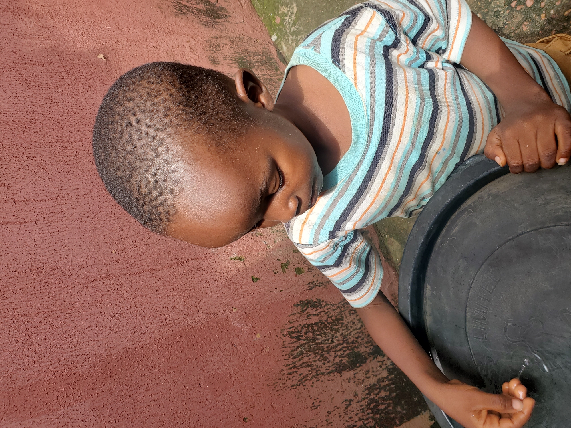 A young child, with dark skin and closely cropped hair, is lying down with their eyes closed, appearing to be sleeping or resting. They are wearing a striped t-shirt with horizontal bands of light blue, white, dark blue, and orange. The child's head is resting on a large, black plastic barrel or container. Water is dripping from the side of the barrel and splashing onto the child's hand, which is positioned on the barrel's rim.
The setting appears to be outdoors, with a reddish-brown ground surface that looks like a paved or concrete area with some texture. There are also patches of green moss or algae visible on the ground and on a concrete structure in the background, suggesting a humid or damp environment. The lighting suggests it is daytime, possibly late morning or afternoon, with sunlight casting shadows. The overall scene conveys a sense of simple, everyday life in a warm climate.
The text "LIMITED" is visible in raised capital letters on the black barrel. The context provided indicates the location is Jos, Nigeria. The child's posture and closed eyes suggest a moment of peaceful repose amidst their surroundings.