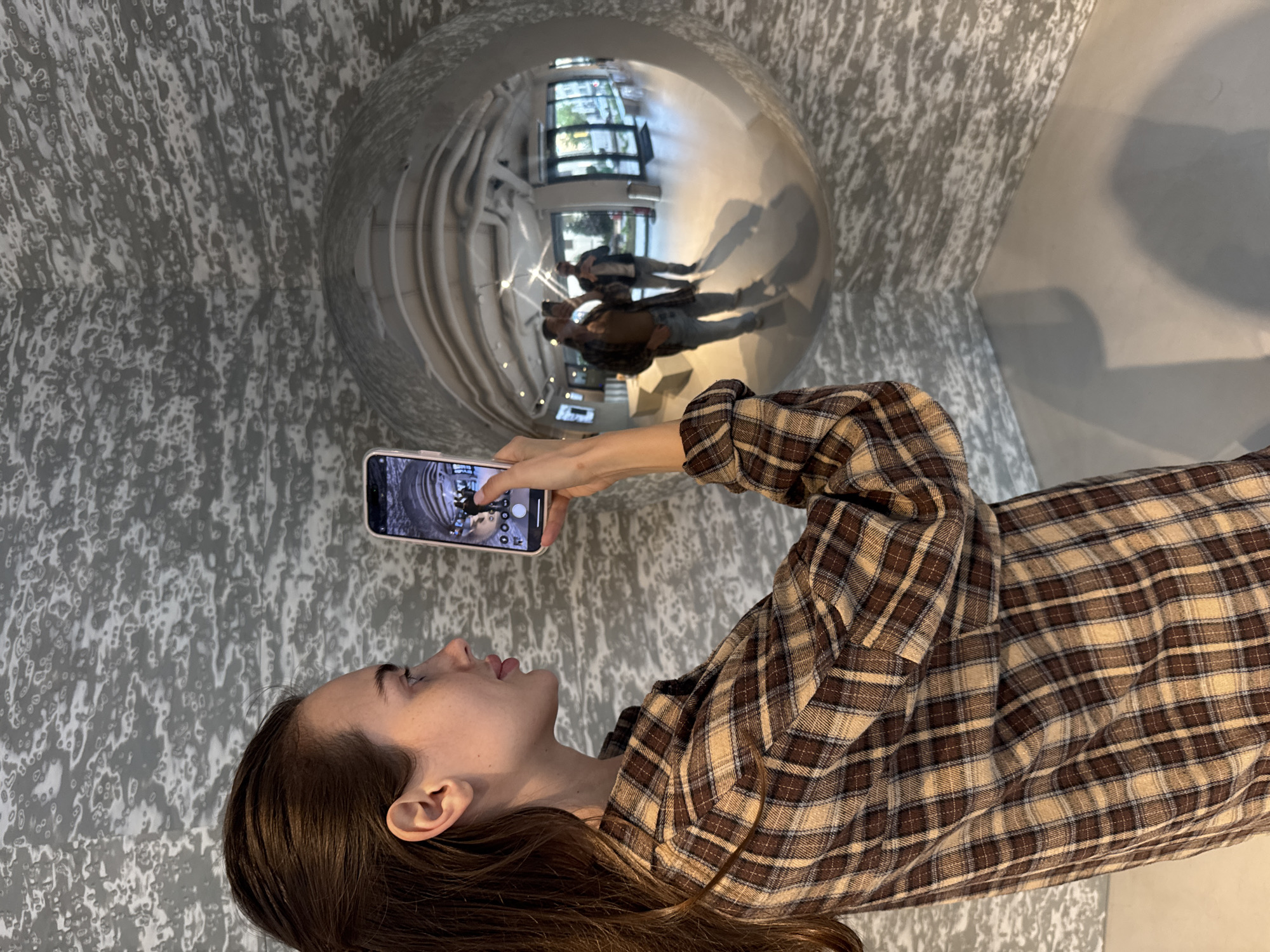 A young woman is captured in a vertically oriented shot, focusing intently on her smartphone screen as she takes a picture. She is seen in profile from the chest up, her dark hair falling down her back. She wears a brown and tan plaid flannel shirt. Her right arm is raised, holding a light-colored smartphone (possibly an iPhone due to its design) horizontally, with the screen facing her and displaying a camera interface.
The subject of her photograph is a large, highly polished silver or chrome spherical object, which occupies a significant portion of the upper left and central area of the image. This sphere acts as a convex mirror, reflecting a distorted view of the interior space. Within the sphere's reflection, the blurred figures of two people are visible; one appears to be the woman taking the photo, and the other, taller figure, is likely a man, both wearing casual clothing (jackets, jeans). Beyond the figures, the reflection shows large windows looking out onto some greenery, and what appear to be industrial-style pipes or ducts on an overhead surface.
The background wall behind the woman and the sphere features a distinctive, mottled gray and white pattern, resembling a textured or abstract design. The lighting suggests an indoor setting with natural light entering from the reflected windows. There is no discernible text visible in the image. The woman appears engaged in documenting the artistic or unusual reflective surface.