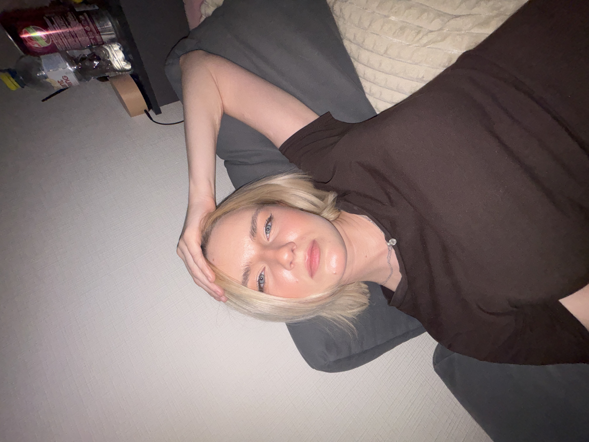 A blonde-haired woman is lying on a gray pillow and what appears to be a patterned mattress or another pillow. She is wearing a brown short-sleeved shirt and a delicate silver necklace with a small charm. Her right arm is bent, with her hand resting on her head. The background includes a gray wall and, in the upper left corner, a shelf with several bottles of beverages. There is no indication of this being Vsevolozhsk, Russia.
