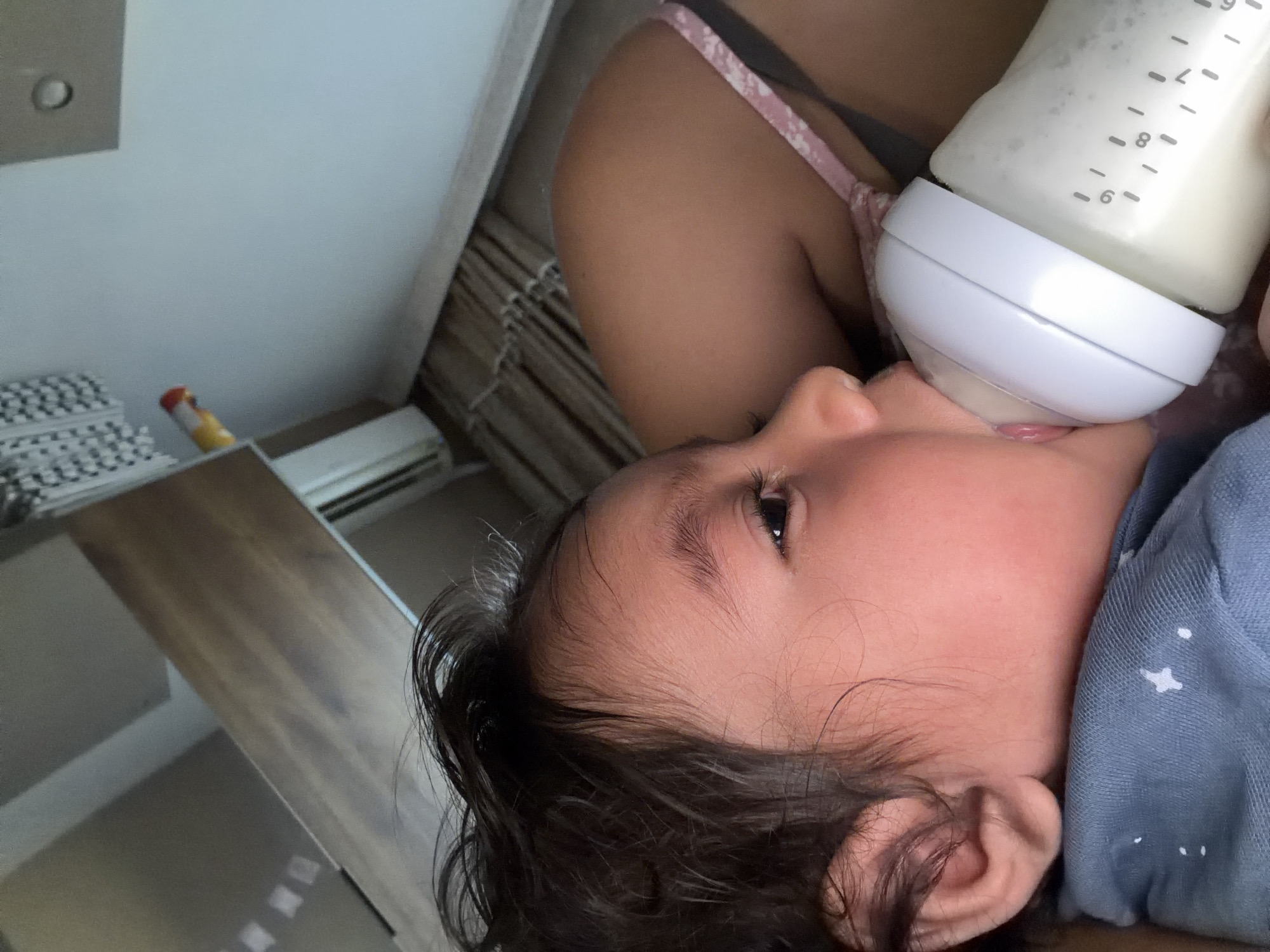 An overhead, close-up shot captures a baby being fed from a bottle. The baby's face, mostly in profile, is turned towards the bottle. The baby has dark hair and a fair complexion. The bottle, held by an unseen person, is a clear plastic baby bottle with white markings indicating measurements. It is filled with a white liquid, presumably milk or formula, and has a white teat. The baby's mouth is open, embracing the teat. The person holding the bottle is partially visible, wearing a pink floral-patterned strap, suggesting a woman. The background consists of blurred interior elements: a light-colored wall on the left, with a grey square fixture; a wooden surface that appears to be a table or counter; and a glimpse of a ribbed texture, possibly blinds or a radiator cover. The overall lighting is soft, suggesting an indoor setting. There is no discernible indication of the location being in Razhevo Konare, Bulgaria.