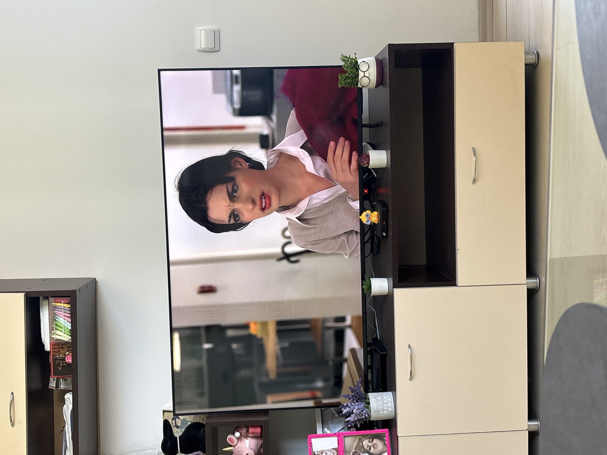 A television shows a woman's face, placed on a dark brown and cream-colored cabinet. A light switch is on the white wall behind the TV. A cabinet is visible to the left, containing various items. The scene is indoors, likely in a home. It is not possible to determine the specific location within the city of Sofia, Bulgaria from the image.