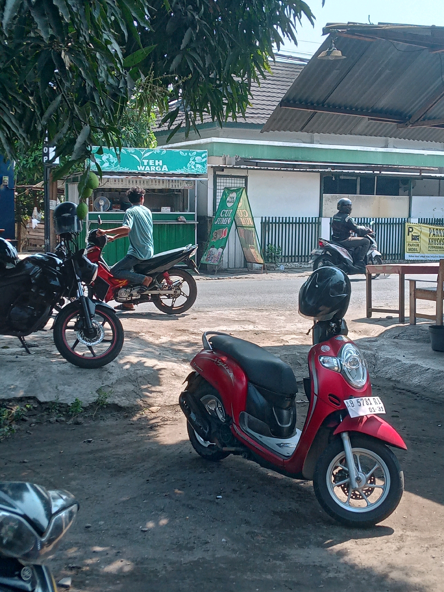 This image captures a street scene in Banguntapan, Indonesia, likely during daylight hours, given the bright sunlight and shadows. In the foreground, a red scooter is parked, its front wheel and part of the body visible. To the left, a portion of a black motorcycle is also visible. Behind these parked vehicles, a man in a blue t-shirt and jeans is seated on a red motorcycle, appearing to be a patron or waiting. Further back, another person wearing a black jacket and helmet is riding a white scooter, moving away from the viewer.
The background reveals a low-rise building with a corrugated metal roof, possibly a shop or food stall. A green sign with white text that reads "TEH WARGA" (meaning "Citizen's Tea") is prominently displayed on the building. Another green banner with text that is partially obscured but includes "CUT" and "JUMBO" is also visible. A white fence runs along the front of the building. To the right, a wooden table and chairs suggest an outdoor seating area. The environment appears to be a somewhat dusty, unpaved roadside with some sparse vegetation. The overall impression is of a casual, everyday scene in a semi-urban Indonesian setting.