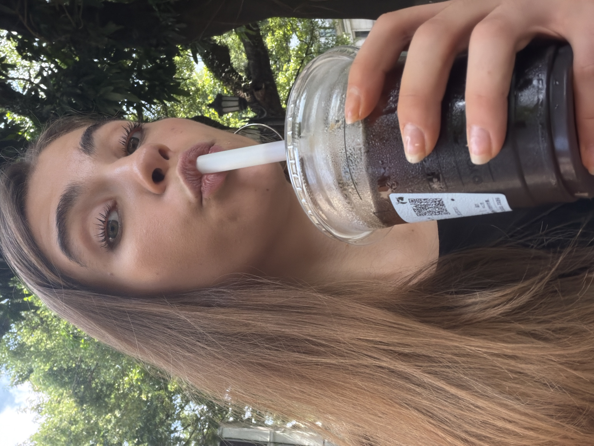 A young Caucasian woman with long, light brown hair is taking a sip of a dark liquid from a clear plastic cup with a white straw. She is holding the cup with her right hand, and her left hand is out of frame. The cup has a QR code sticker on it, and the liquid appears to be iced coffee or a similar beverage. The woman is outdoors, with trees and greenery in the background. A street lamp is visible in the upper right quadrant of the image. The image is a close-up shot, focusing on the woman's face and the cup. It is unclear if the location is Liwan District, China, as there are no specific visual cues to confirm this.