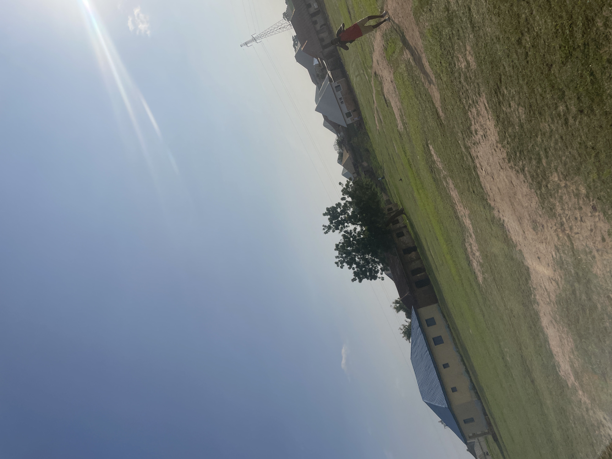 The image depicts a clear sky with a sun glare in the upper right quadrant. In the foreground, a person in a red shirt and beige shorts is standing in a grassy field. The field is mostly green with patches of dirt. In the middle ground, several buildings with distinct architectural styles are visible. One building in the left middle ground has a light yellow facade and a blue metal roof with a distinctive geometric shape. Further back, a line of buildings with various roof types, including what appears to be corrugated metal and tiled roofs, stretches across the scene. A large tree is situated between the yellow building and the row of houses. In the background, a tall, lattice-style electricity pylon is visible against the sky. The overall setting suggests a residential or community area in Keffi, Nigeria, with elements of modern infrastructure present.