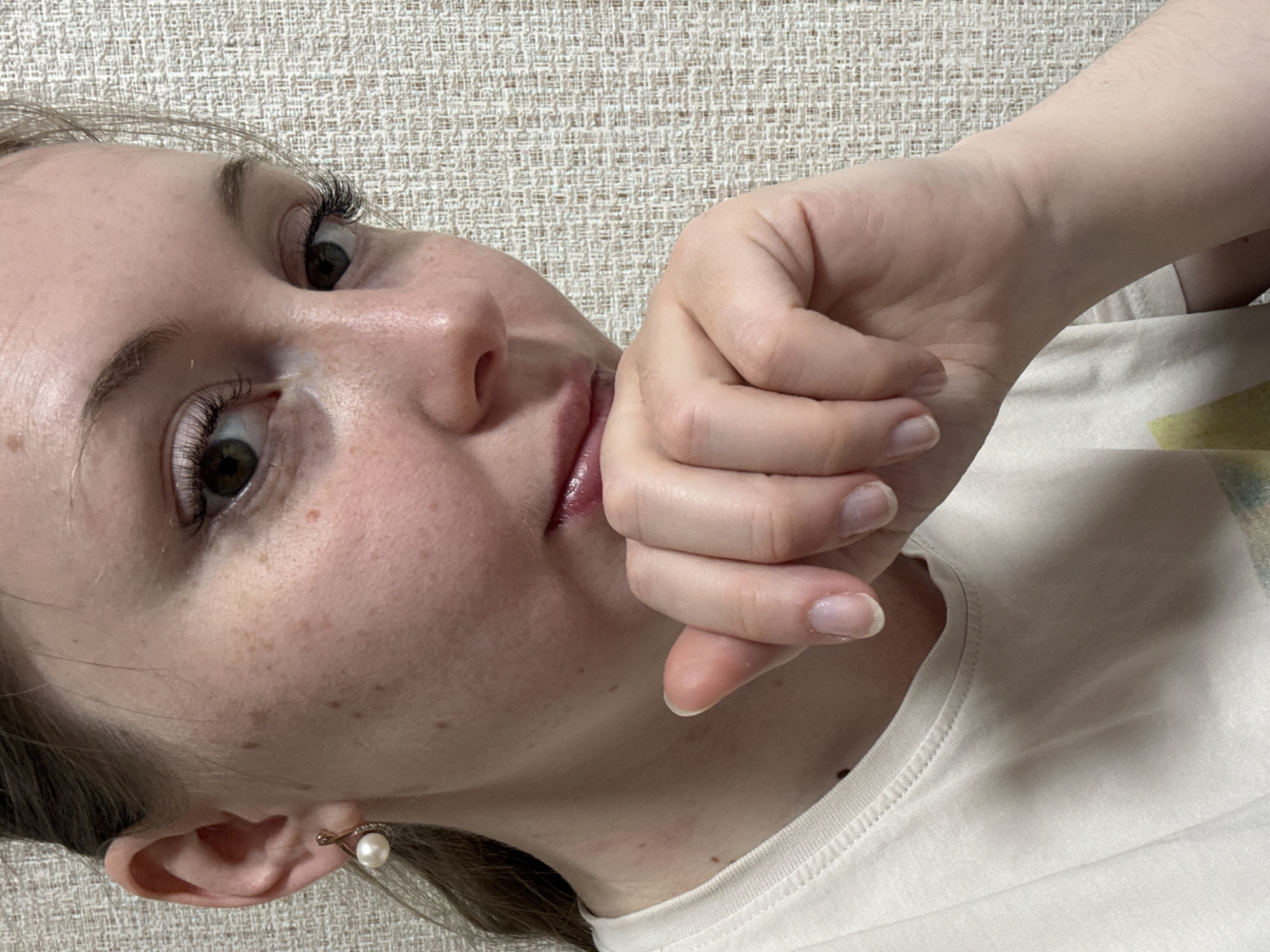 A close-up, slightly angled view shows a fair-skinned person with light brown hair. The person's face is prominent, freckled, and features brown eyes with extended eyelashes. Their left ear is visible, adorned with a pearl earring. The person's right hand is brought up towards their face, with the fingers curled towards the mouth. They wear a light-colored t-shirt. The background consists of a textured neutral-toned surface. The location is Unknown, Unknown.