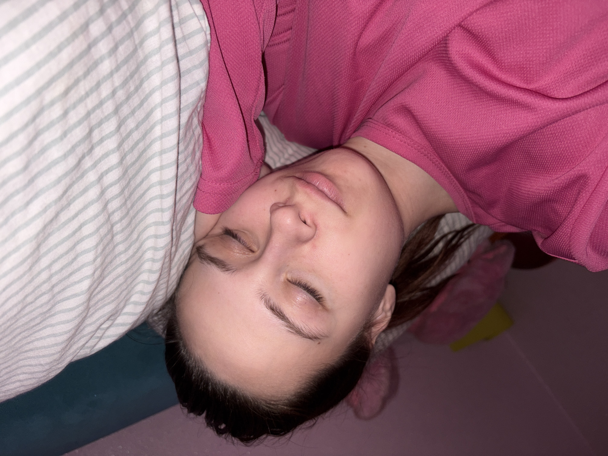 A person is lying down with their eyes closed. They are wearing a pink shirt and resting on a pillow with gray and white stripes. The surroundings are dimly lit, with a pinkish hue dominating the background. The location of the image is not discernable from the provided picture.
