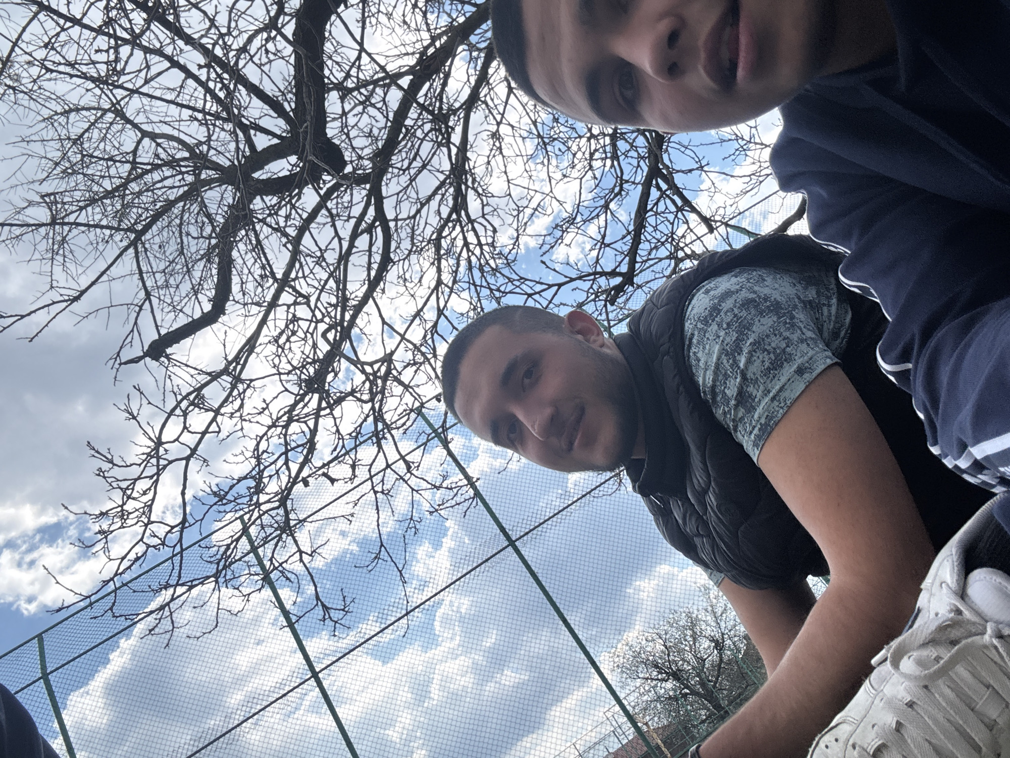Two young men are photographed from a low angle against a backdrop of a bare tree, a wire fence, and a cloudy sky. The image appears to have been taken outside, possibly in a park or recreational area, with glimpses of greenery and buildings in the background. The men are positioned in the upper right of the frame. One is wearing a navy jacket and the other a black vest over a patterned shirt. White sneakers are visible at the bottom. The photograph was taken in Topoli, Bulgaria.