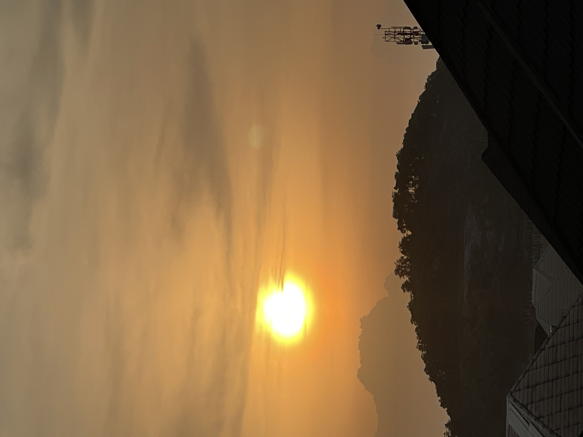 This is a vertical, medium-shot photograph taken at sunset in Abuja, Nigeria. The sun, a bright, luminous orb, is positioned in the upper left quadrant of the image, casting a warm, orange glow across the sky. Wispy clouds are scattered throughout the sky, adding texture and depth. In the mid-ground, a dark, silhouetted hill rises, topped with a line of trees. In the foreground, the dark, angular shapes of rooftops are visible, suggesting a residential area. A cell tower stands tall on the right side of the image, silhouetted against the sky. The overall mood of the image is serene and tranquil, capturing the beauty of a sunset over the cityscape.