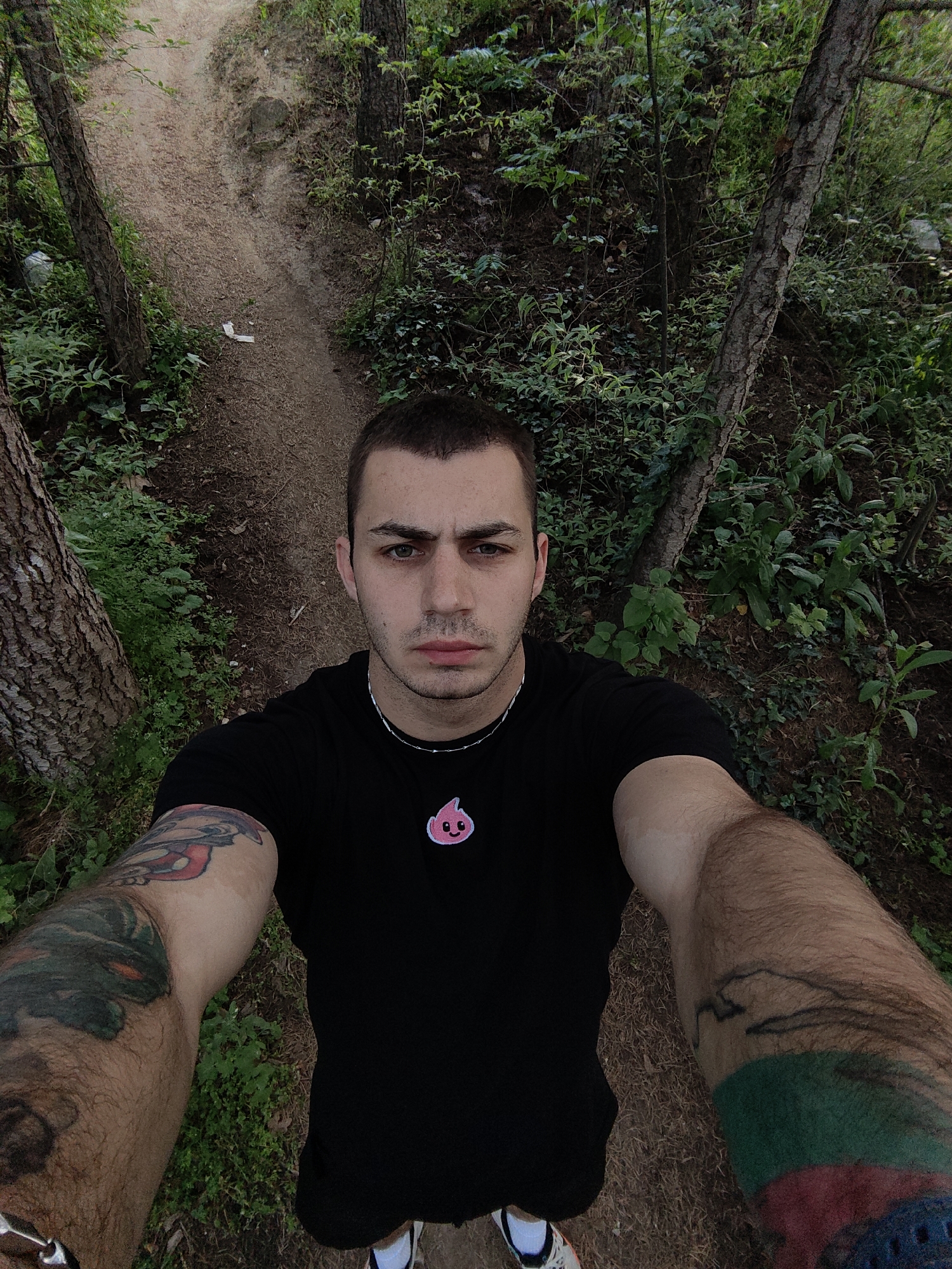 A man takes a selfie on a dirt path in a wooded area. The path leads uphill away from the man. The man is wearing a black t-shirt with a small pink cartoon emblem on the chest, a silver necklace, and athletic shoes. He has tattoos on both forearms. The environment is green with trees and undergrowth surrounding the path. It's possibly located within the Varna area, given the dense woodlands common around the city.