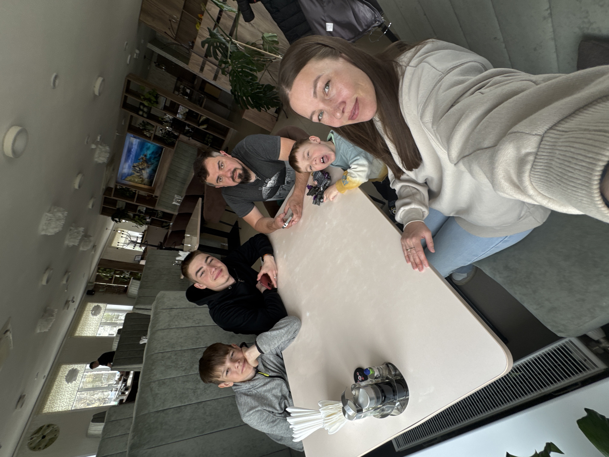 The image shows a family of five posing for a selfie at a table. The setting appears to be inside a restaurant or cafe. The location is the city of Chaikovsky, Russia.
The selfie is taken by an adult woman wearing a light-colored hoodie and jeans. Standing next to her is an adult man in a gray t-shirt. A young child is held between them, making a face for the camera while holding a toy. At the opposite end of the table are two other young boys, dressed in casual clothing. One wears a black hoodie, and the other wears a gray hooded sweatshirt.
On the table, there is a condiment holder with a napkin dispenser. The table itself is light-colored and has a rounded rectangular shape. Beyond the table, there is upholstered seating and a radiator. In the background, there are windows, a display shelf, and interior decor. The environment indicates an interior setting with artificial lighting.