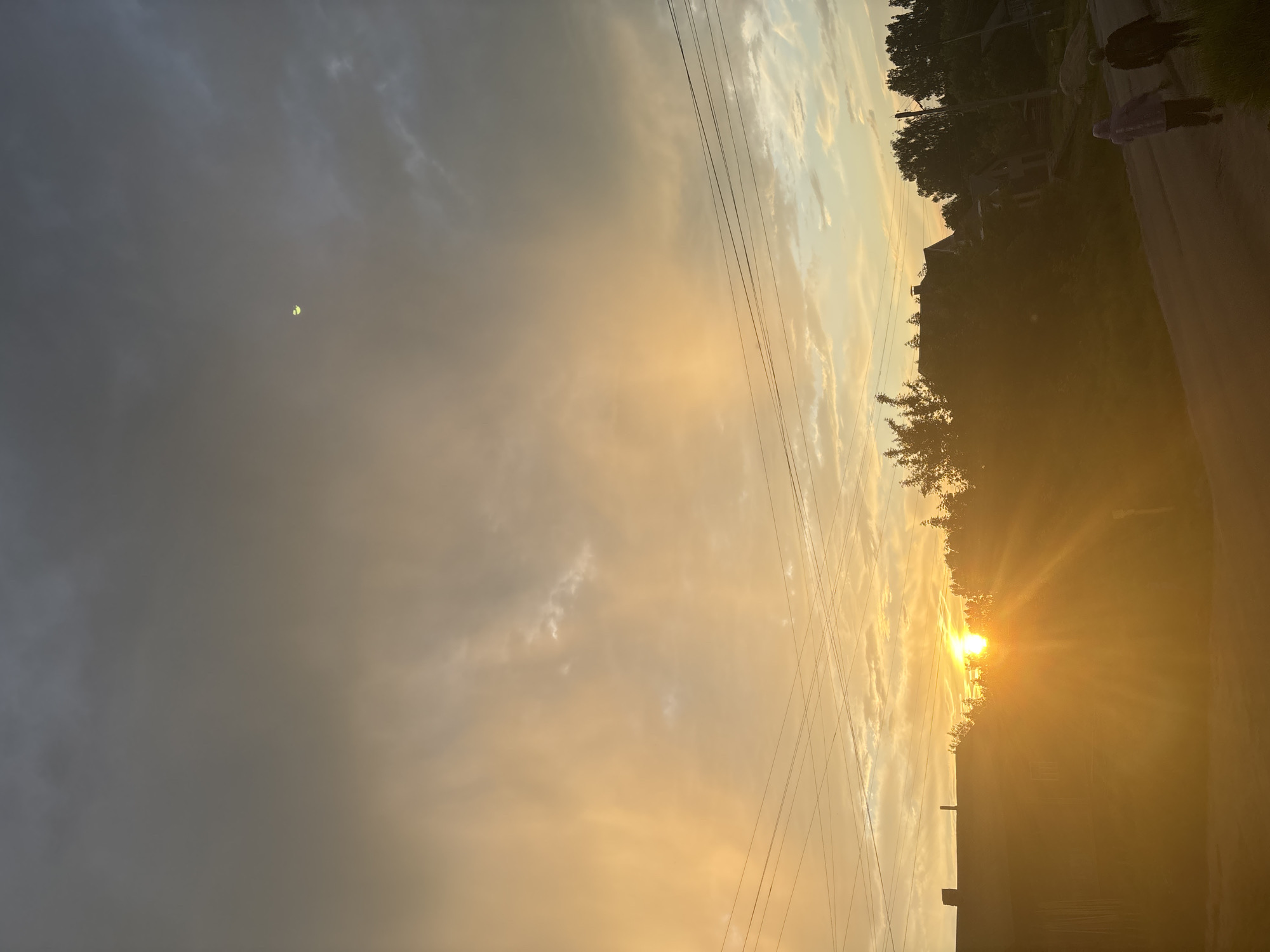 The image depicts a sunset over a village, likely in Krasnye Baki, Russia. The sky is filled with dramatic clouds, transitioning from dark grey at the top to warm orange and yellow hues near the horizon. A crescent moon is faintly visible in the upper right portion of the sky. Several power lines stretch across the frame, silhouetted against the bright sky. In the foreground, the silhouette of a wooden house is visible on the left. Trees and the roofs of other houses are scattered throughout the middle ground, partially obscured by the low-angled sunlight. On a paved road in the lower right, two figures, appearing to be people, are walking away from the viewer, heading towards the right side of the frame. The overall atmosphere is serene and evocative of a rural evening.