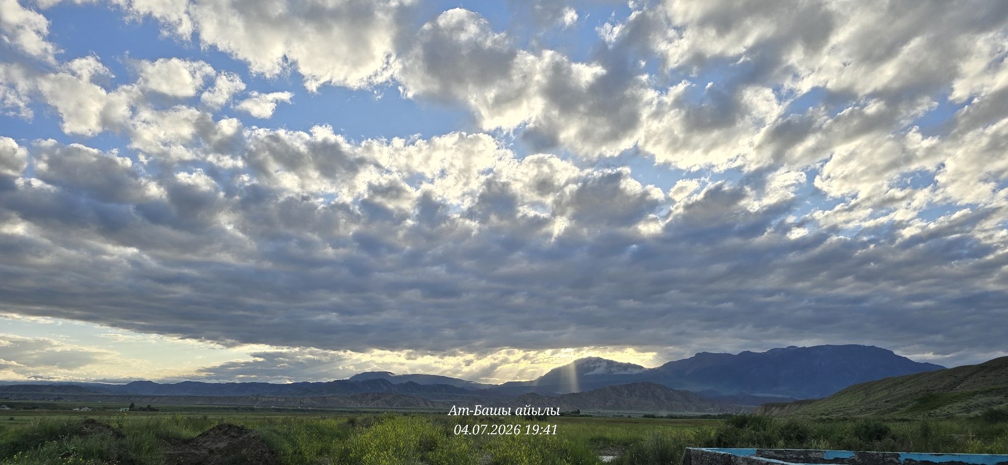 The image captures a vast sky filled with a dramatic pattern of clouds, ranging from bright white to dark gray, illuminated by sunlight that breaks through in rays. Below the expansive sky, a landscape unfolds featuring rolling hills and distant mountains. The foreground is characterized by green grass and some disturbed earth. In the mid-ground, a flat expanse of land is visible with scattered structures, suggesting a rural or semi-rural area. The text overlay indicates the location as "At-Bashy, Kyrgyzstan" and the date and time as "04.07.2026 19:41." The scene appears to be a natural landscape photograph taken during the late afternoon or early evening, given the lighting conditions.