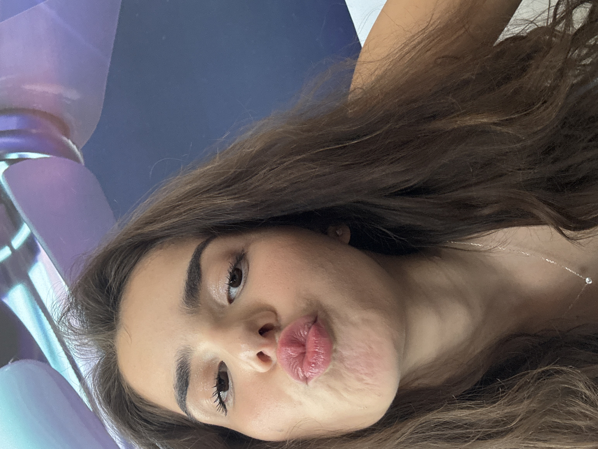 The image shows a close-up, selfie-style shot of a young woman with long, brown hair, puckering her lips. She is wearing light makeup, including mascara and what appears to be lip gloss. A delicate silver necklace with a small pendant is visible around her neck. The background is out of focus and appears to be an indoor setting with a blue wall and some abstract, possibly artistic, elements in light colors at the top of the frame. There are no discernible elements in the image that would indicate a specific location within the city of Shumen, Bulgaria.