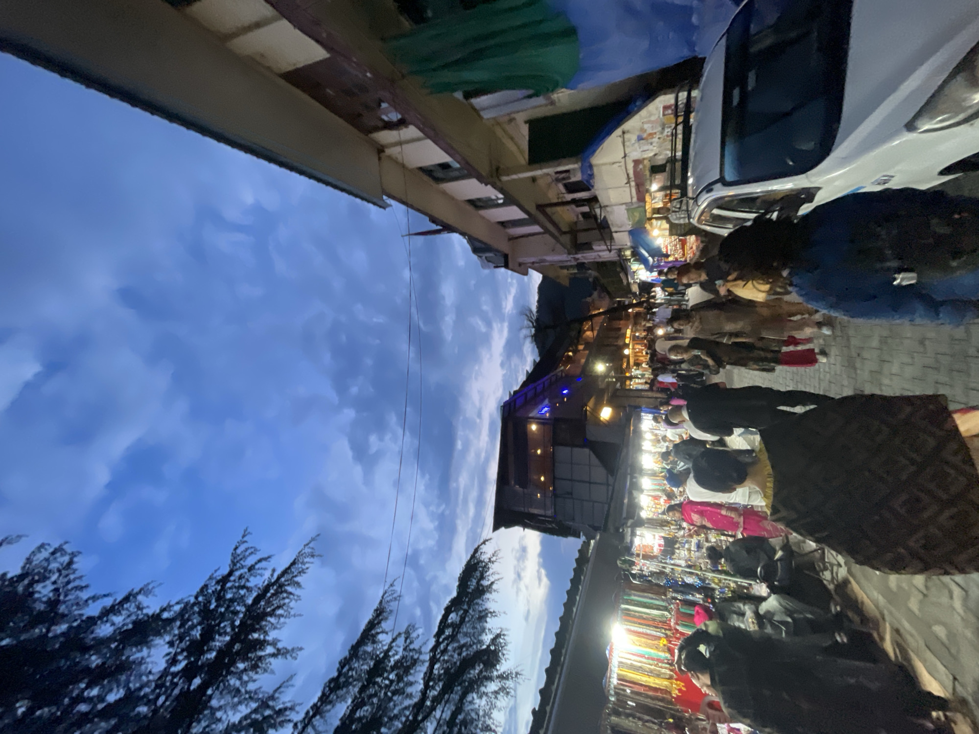 The image depicts a street scene in McLeod Ganj, a town in Dharamshala, India, during twilight. A crowd of people is walking along a bustling street lined with shops and market stalls. The shops on the left display colorful textiles and jewelry, illuminated by bright lights, contrasting with the darkening sky above. On the right, a multi-story building with balconies overlooks the street, and a white taxi is parked. The sky is a deep blue with visible clouds, suggesting the evening is setting in. The overall atmosphere appears lively and commercial, with a mix of locals and possibly tourists browsing the shops.