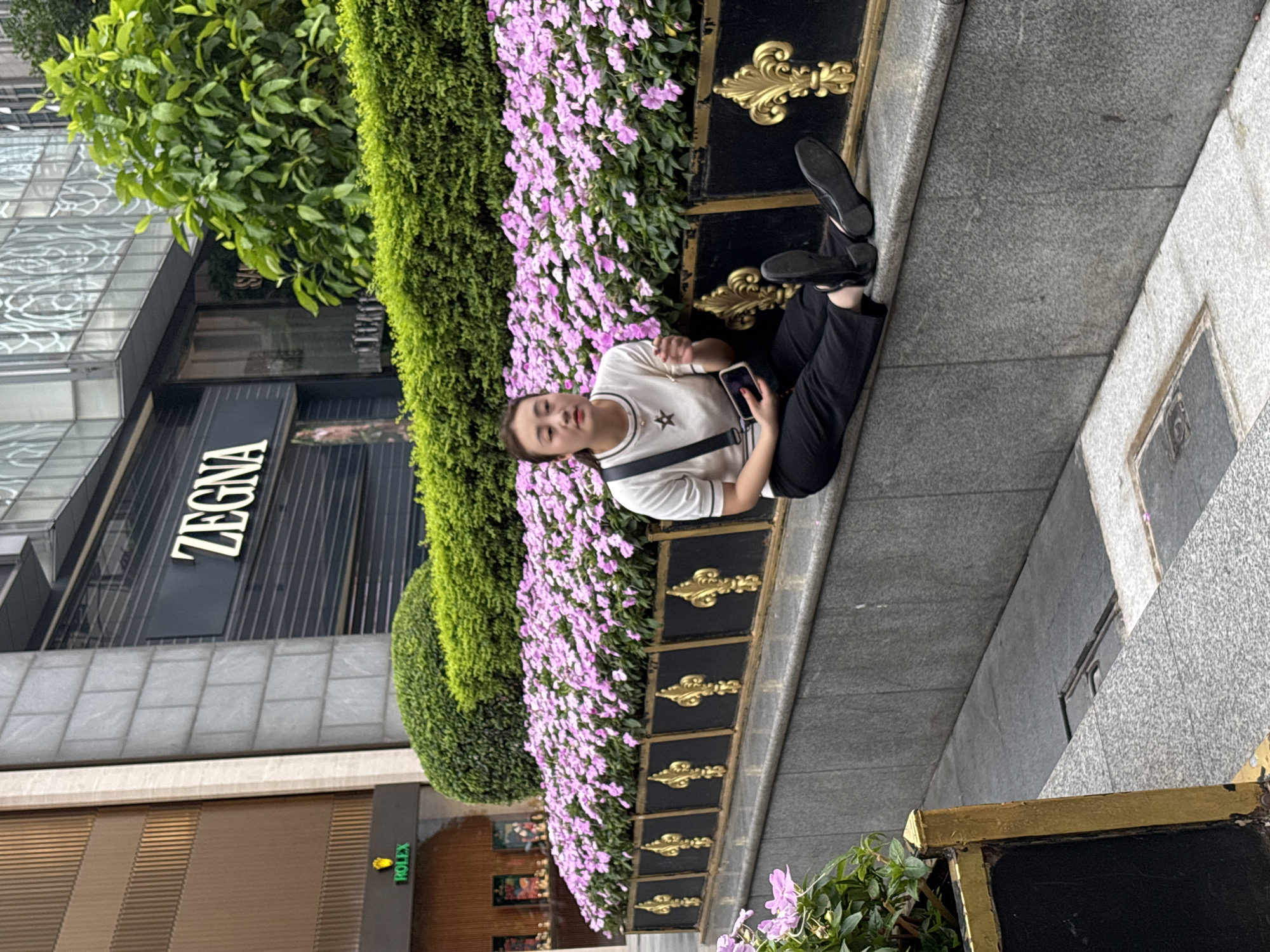 A person sits with their legs extended to the right on a gray sidewalk next to a long planter box filled with light purple flowers and a tall green hedge. The planter box is decorated with gold accents. The person wears black pants, a white t-shirt with a dark star design, and black shoes, and they hold a cell phone in their right hand. Behind the planter box is a building with signs for Zegna and Rolex. The scene is in Yuzhong District, China.