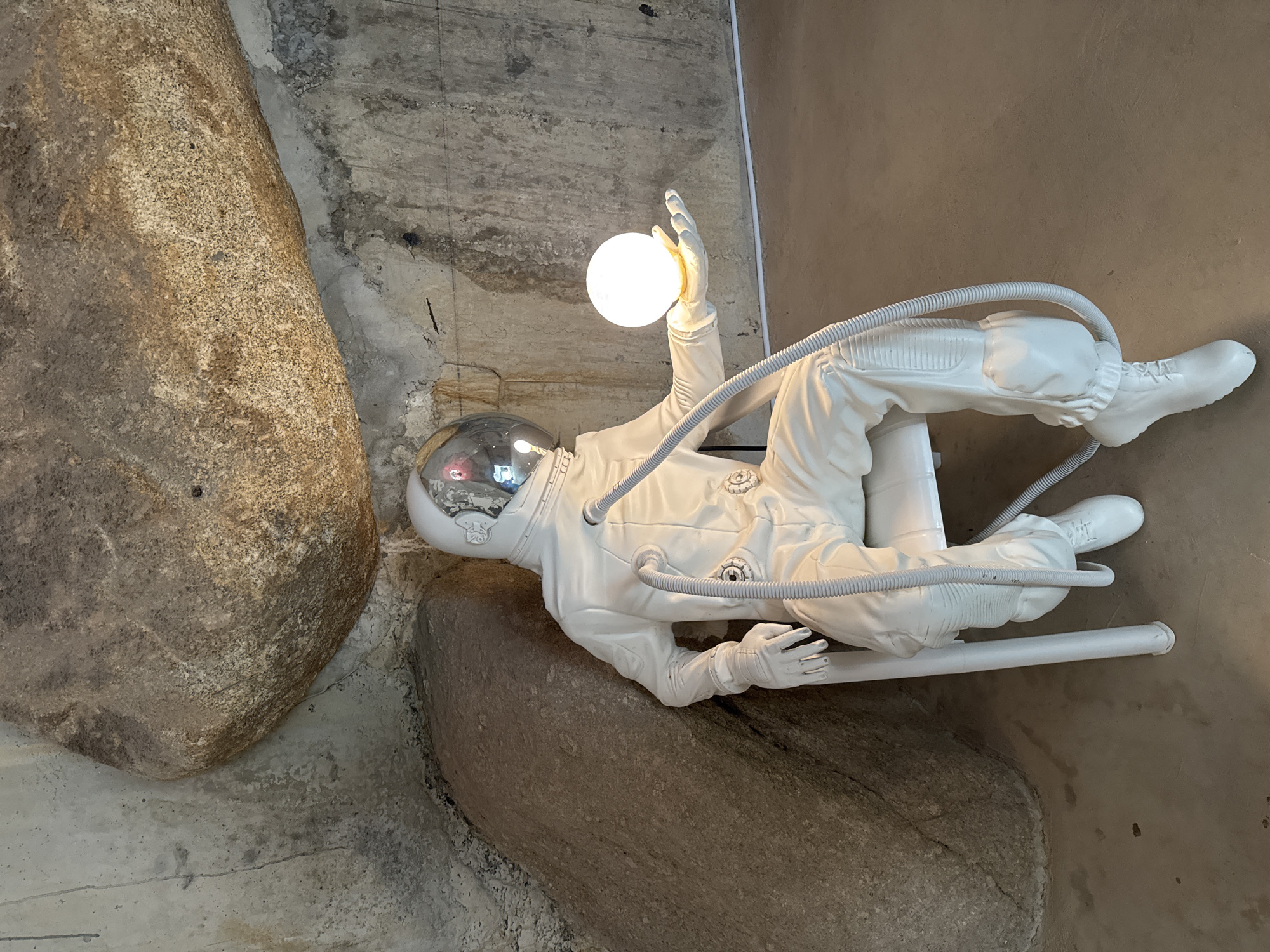 The image shows a white astronaut statue lying horizontally against a concrete wall with two large rocks. The statue is wearing a spacesuit with a helmet and has one arm raised, holding a glowing white orb. The statue appears to be supported by a white pole. The location is Baan Lipa Noi, Thailand.