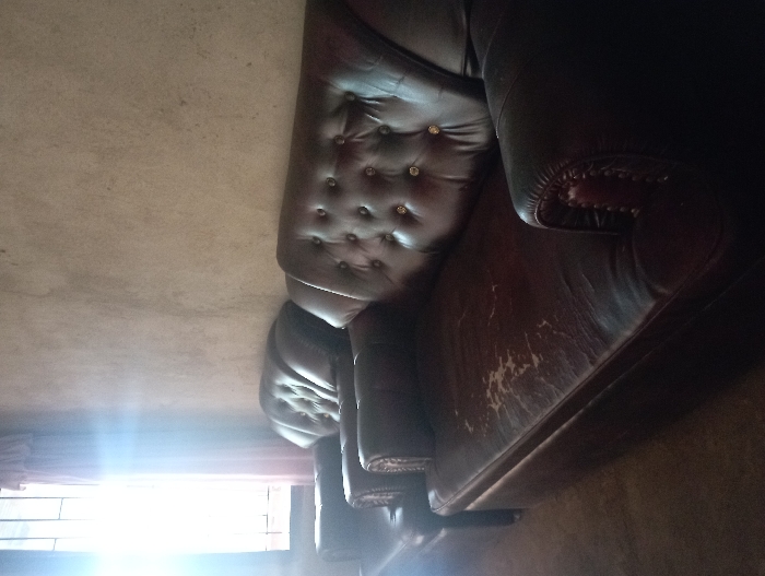 This media file captures an indoor scene, primarily featuring a dark brown leather sofa. The sofa is designed in a Chesterfield style, identifiable by its deeply tufted backrest adorned with numerous buttons, some appearing to be gold-colored. A classic rolled armrest is visible on the right side of the frame.
The leather upholstery on the front and seating area of the sofa shows significant signs of wear and tear, with numerous scuff marks and lighter patches where the finish has degraded. The overall impression is of an aged but robust piece of furniture.
A bright light source, likely a window, is positioned on the left side of the image, causing a strong glare that illuminates part of the room and the lower portion of the sofa. The background on the upper left appears to be a light-colored, possibly textured wall or floor surface. No people or activities are depicted in the image. There is no visible text. The context provided indicates this scene is located in Pubian, Indonesia.