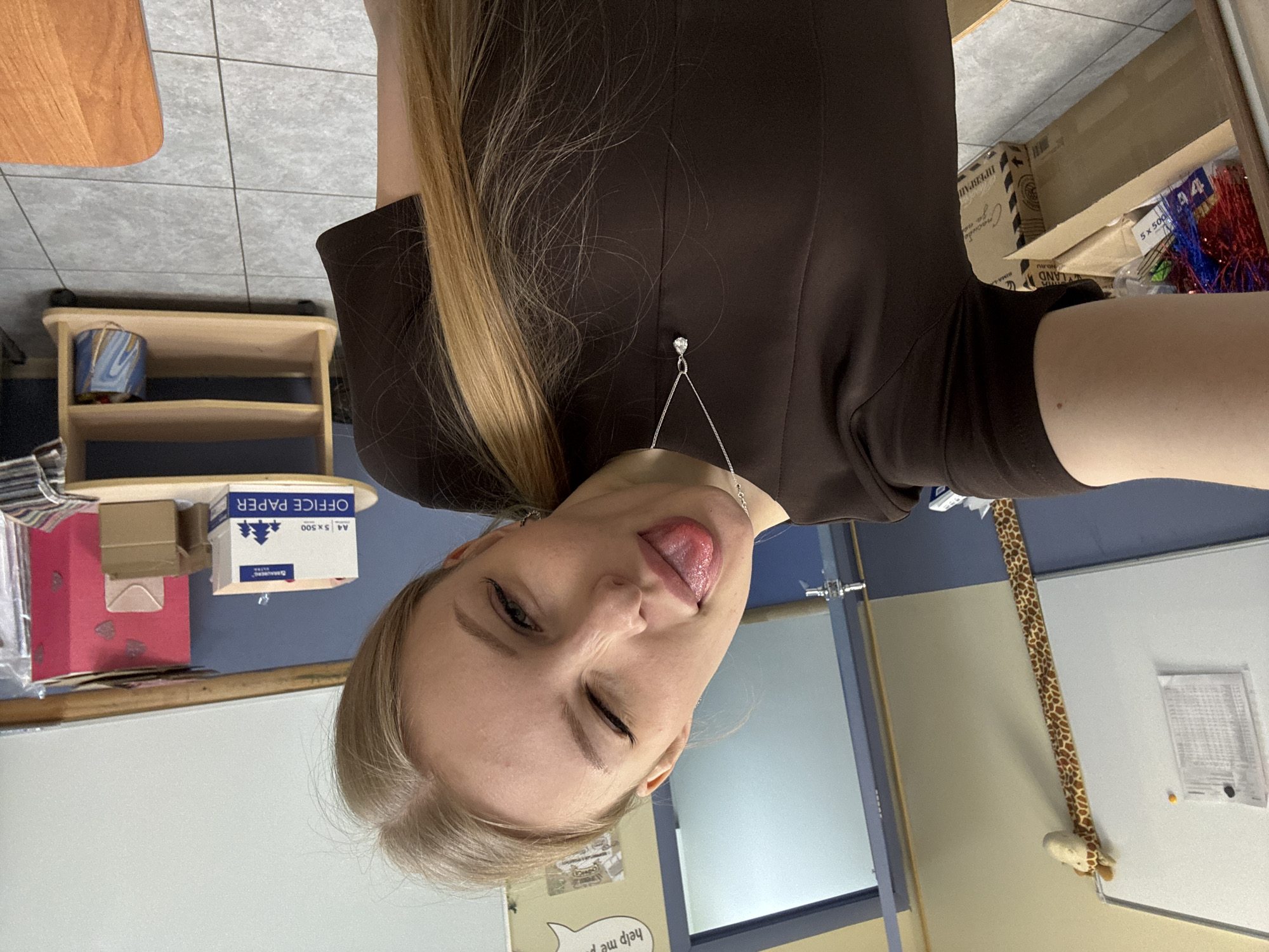A woman is taking a selfie. She has fair skin, blonde hair, and is wearing a brown top and a silver pendant necklace. She is sticking her tongue out and winking. She is in what appears to be an office or school environment. To the left of the image is a small wooden shelf, a pink gift bag, and a box labeled "A4 5 x 500 OFFICE PAPER". To the right, there is a partial view of a box labeled "ПРЕВРАП" in Russian, along with other items. There is a door and a wall with a stuffed giraffe hanging on it. There is also a speech bubble containing the words "help me p". There is no indication of a city.