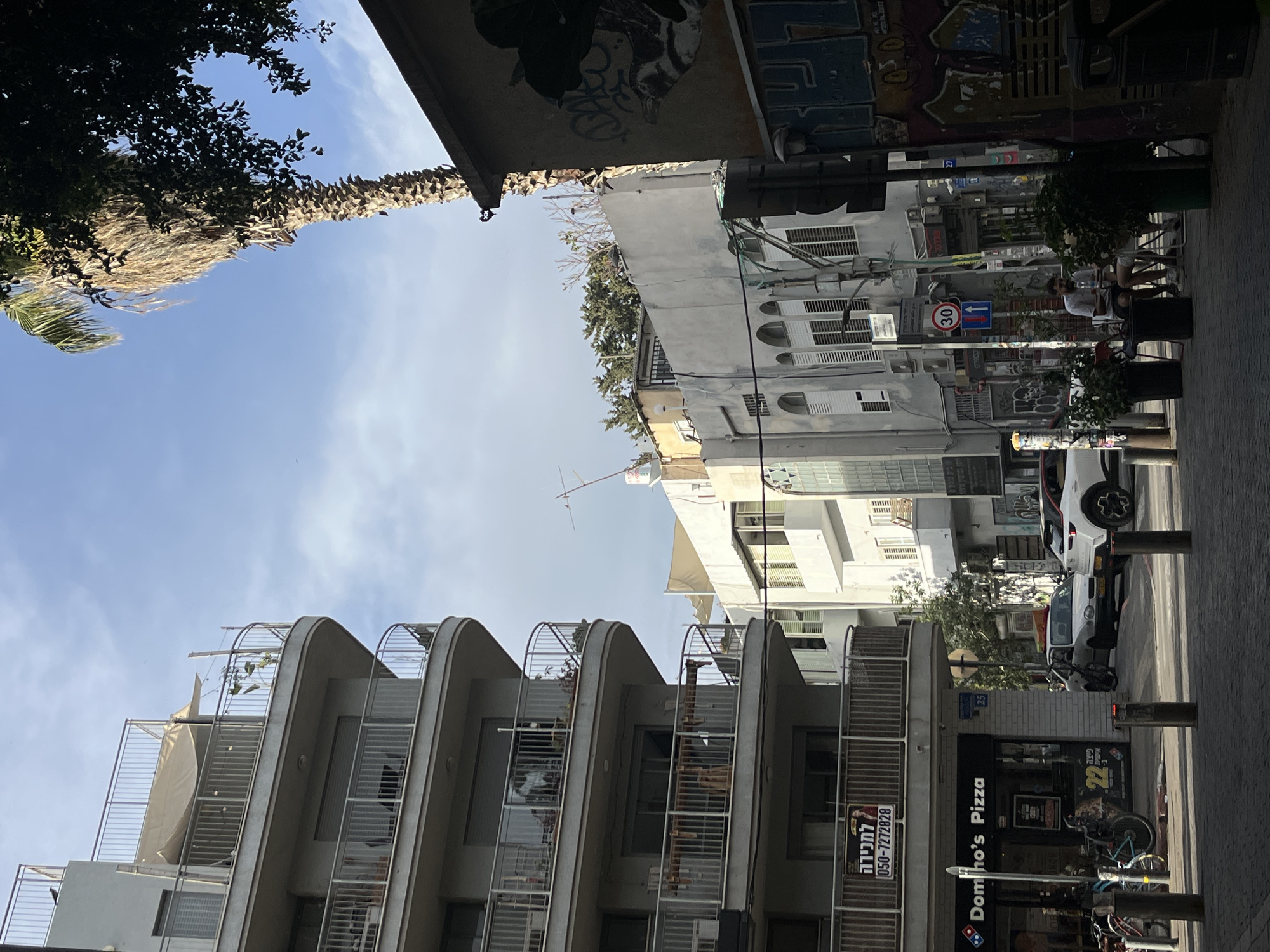 The image depicts a street view in Tel Aviv, Israel. The buildings are a mix of modern and older styles, with white or gray facades. A curved building with balconies is visible on the lower left. There's a Domino's Pizza store at street level, with a sign visible. Street signs and parked cars are also seen. The sky is blue with light clouds. Palm trees and other greenery are visible, adding to the urban landscape. People are sitting at outdoor cafes, indicative of street life. Graffiti is visible on some buildings.