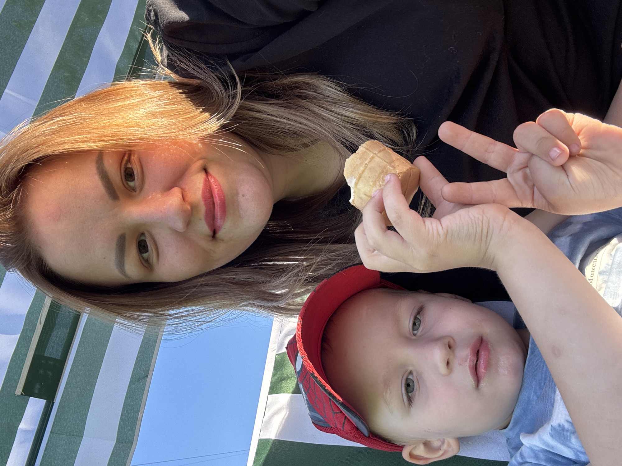A close-up, selfie-style shot shows a woman and a young boy outdoors, seated under a striped awning. The woman, on the right, has light brown hair and is wearing a black top. Her right hand is holding a piece of an ice cream cone to her mouth, and her left hand is held up with two fingers extended in a peace sign. The boy, on the left, is wearing a red baseball cap with a Spiderman design and a blue tie-dye t-shirt. He is looking towards the camera with his mouth slightly open. The background consists of a blue sky and the green and white striped awning. The scene appears to be a casual outdoor setting, possibly a park or backyard, in the city of Chaikovsky, Russia.